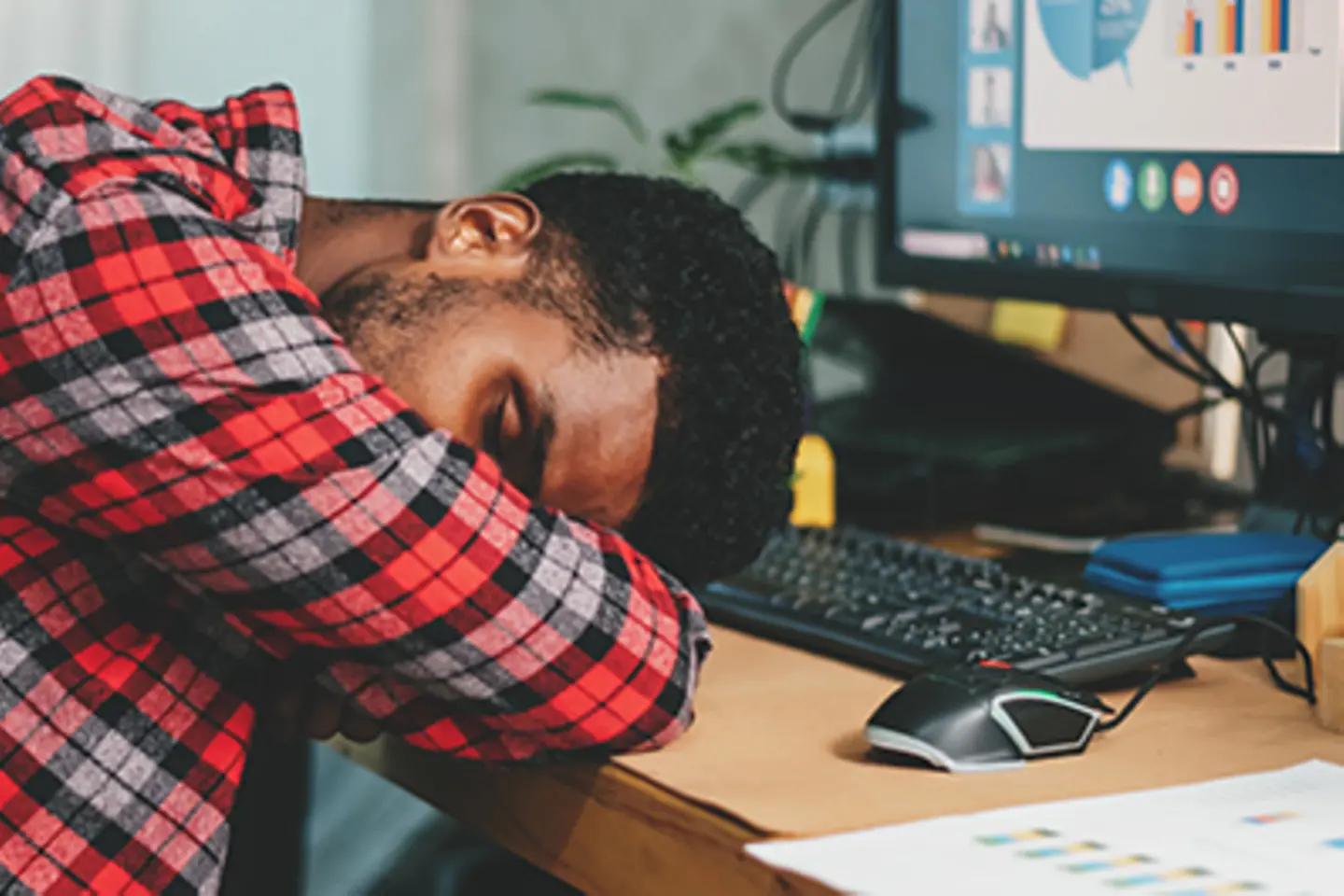 ein Mann sitzt vor einem Bildschirm und hat den Kopf mit verschlossenen Augen auf seine verschränkten Arme gelegt