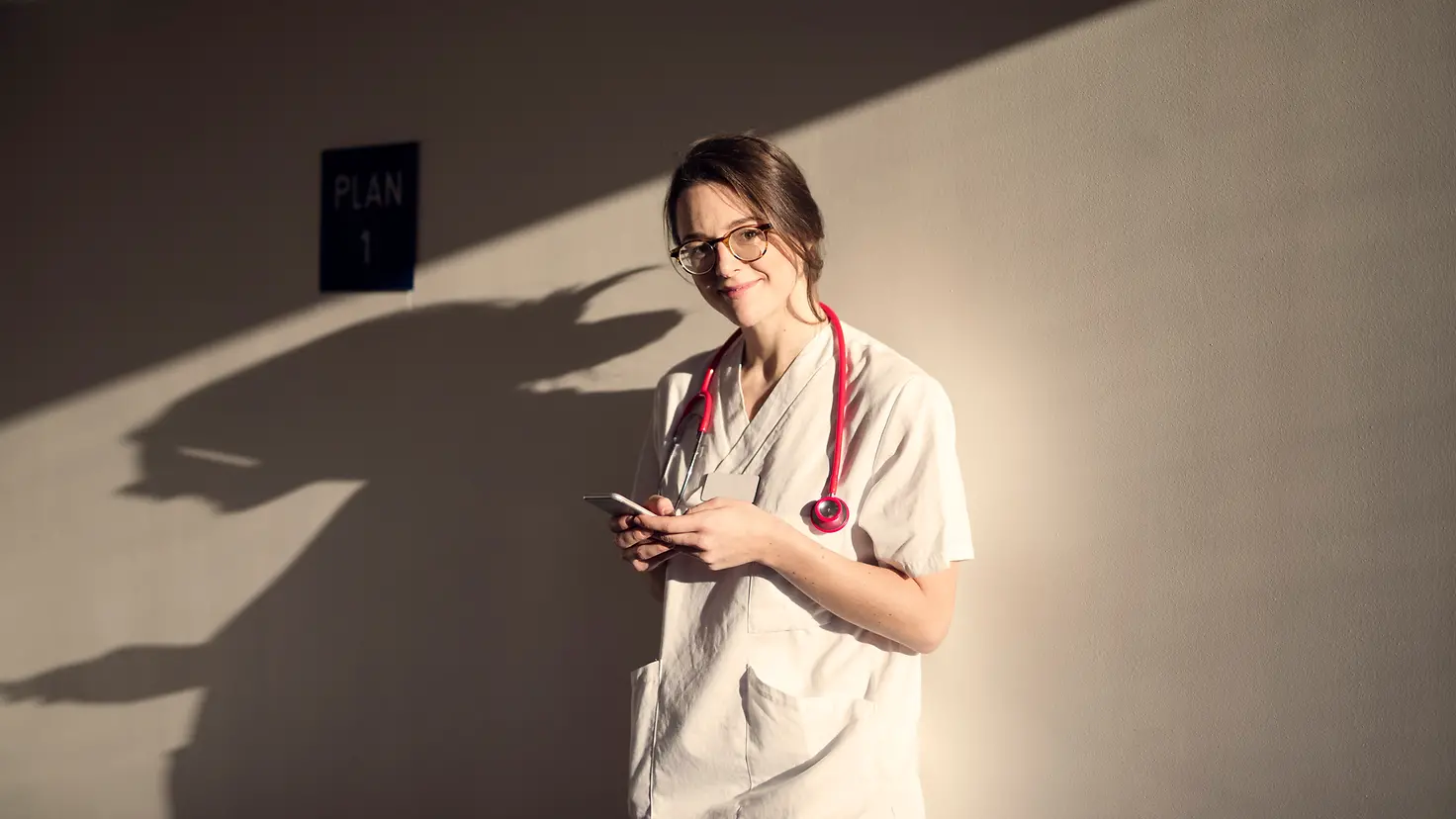 Eine Ärztin steht vor einer Wand, hält ihr Smartphone in der Hand und schaut lächelnd in die Kamera.