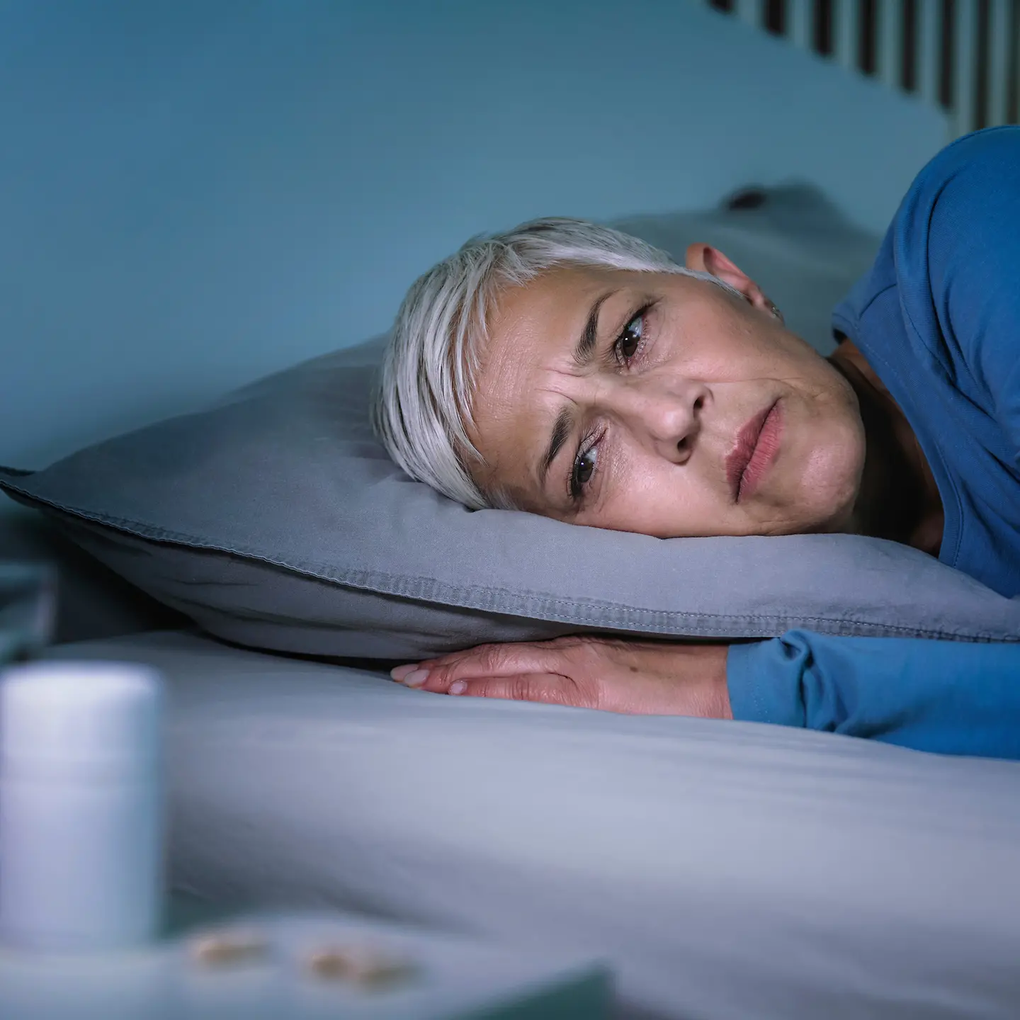 Zu sehen ist eine Frau, die wach im Bett liegt und offensichtlich nicht schlafen kann. Auf dem Nachttisch vor ihr steht ein Glas Wasser und liegen Tabletten.