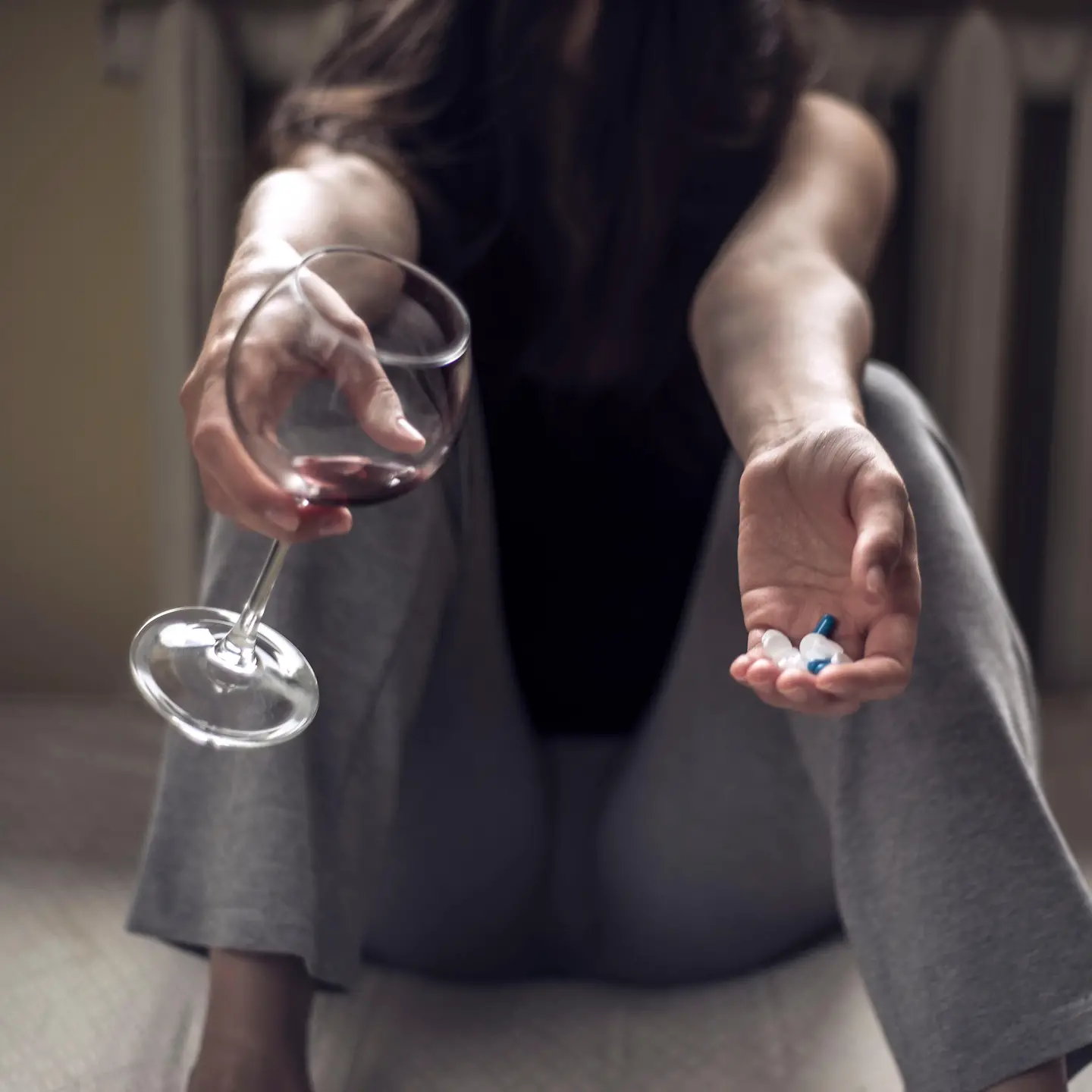 Zu sehen ist eine sitzende Frau, mit einem Rotweinglas in der einen und Pillen in der anderen Hand