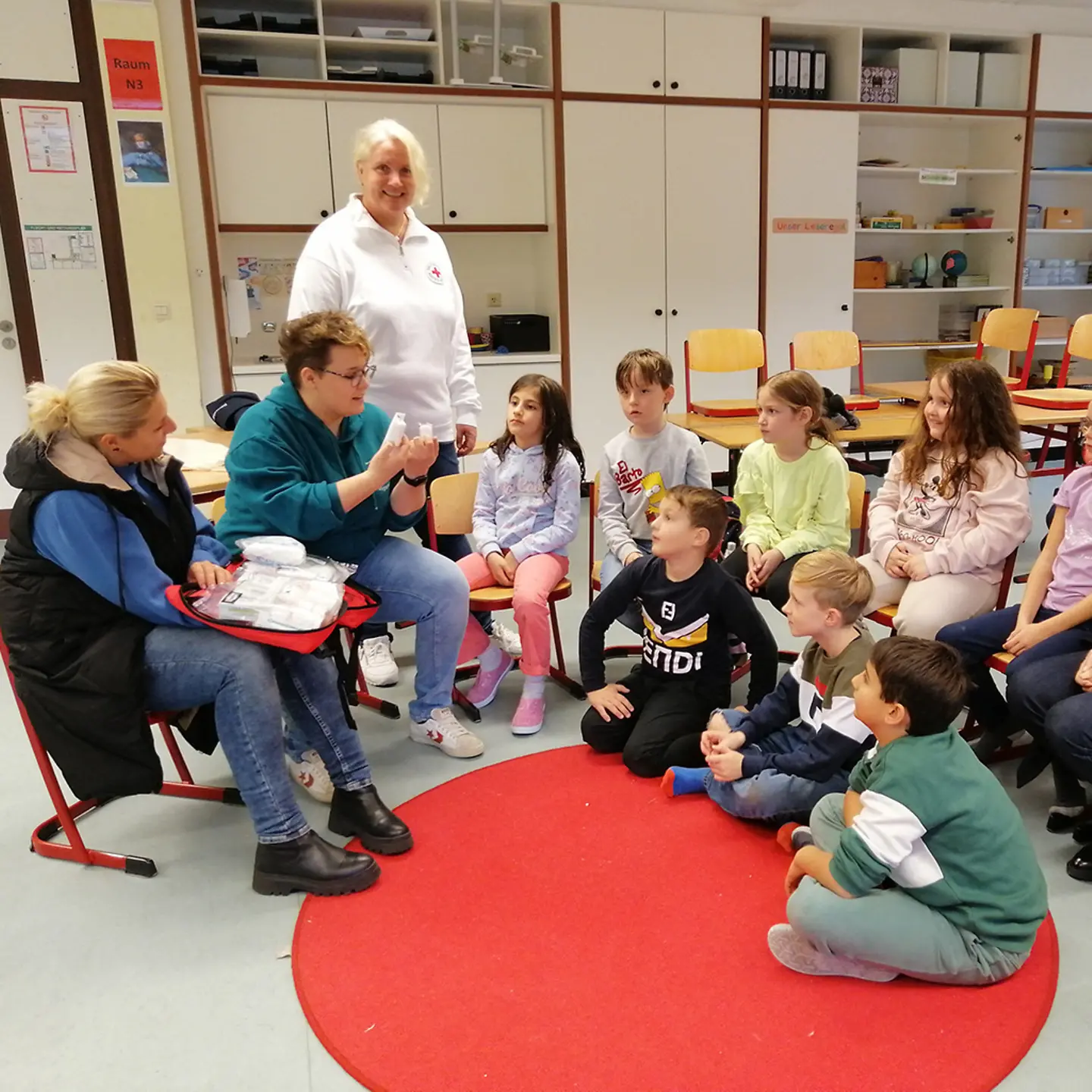 Stuhlkreis. Eine Frau zeigt einer Gruppe Jungen und Mädchen, wie sie einen Druckverband anlegen.