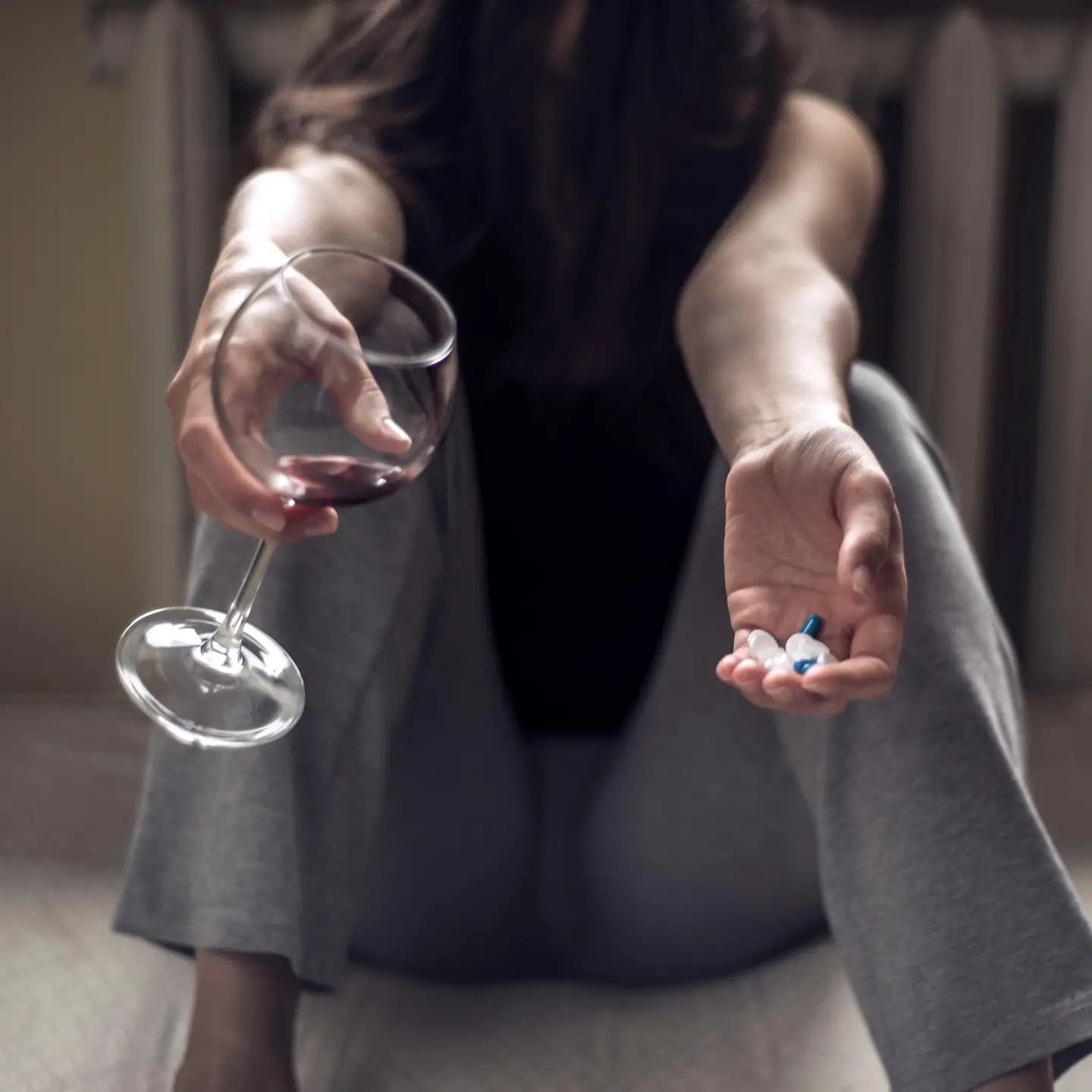 Zu sehen ist eine sitzende Frau, mit einem Rotweinglas in der einen und Pillen in der anderen Hand