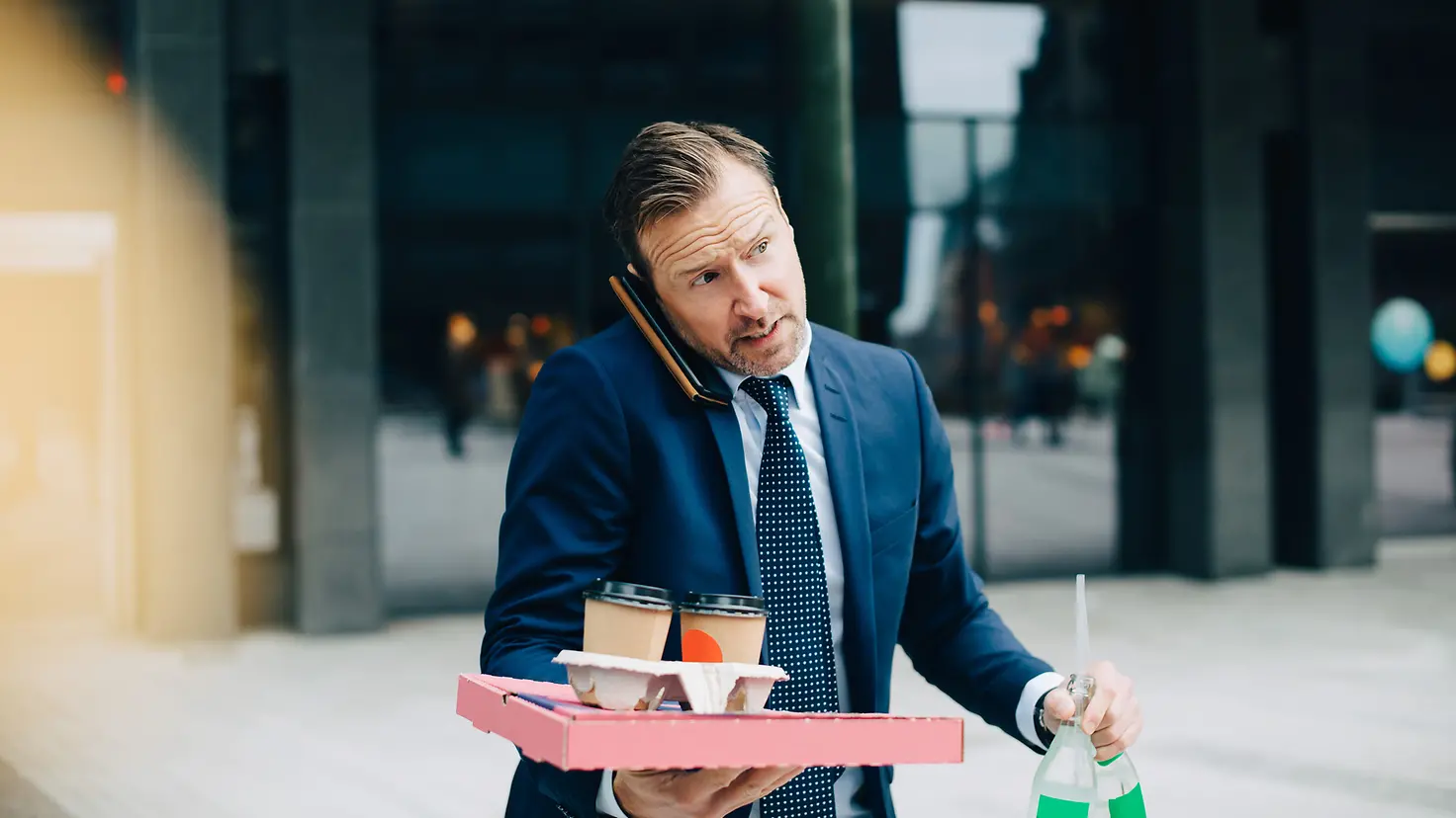 Geschäftsmann steht unter Leistungsdruck und telefoniert unterwegs mit Kaffee und Getränken in den Händen.