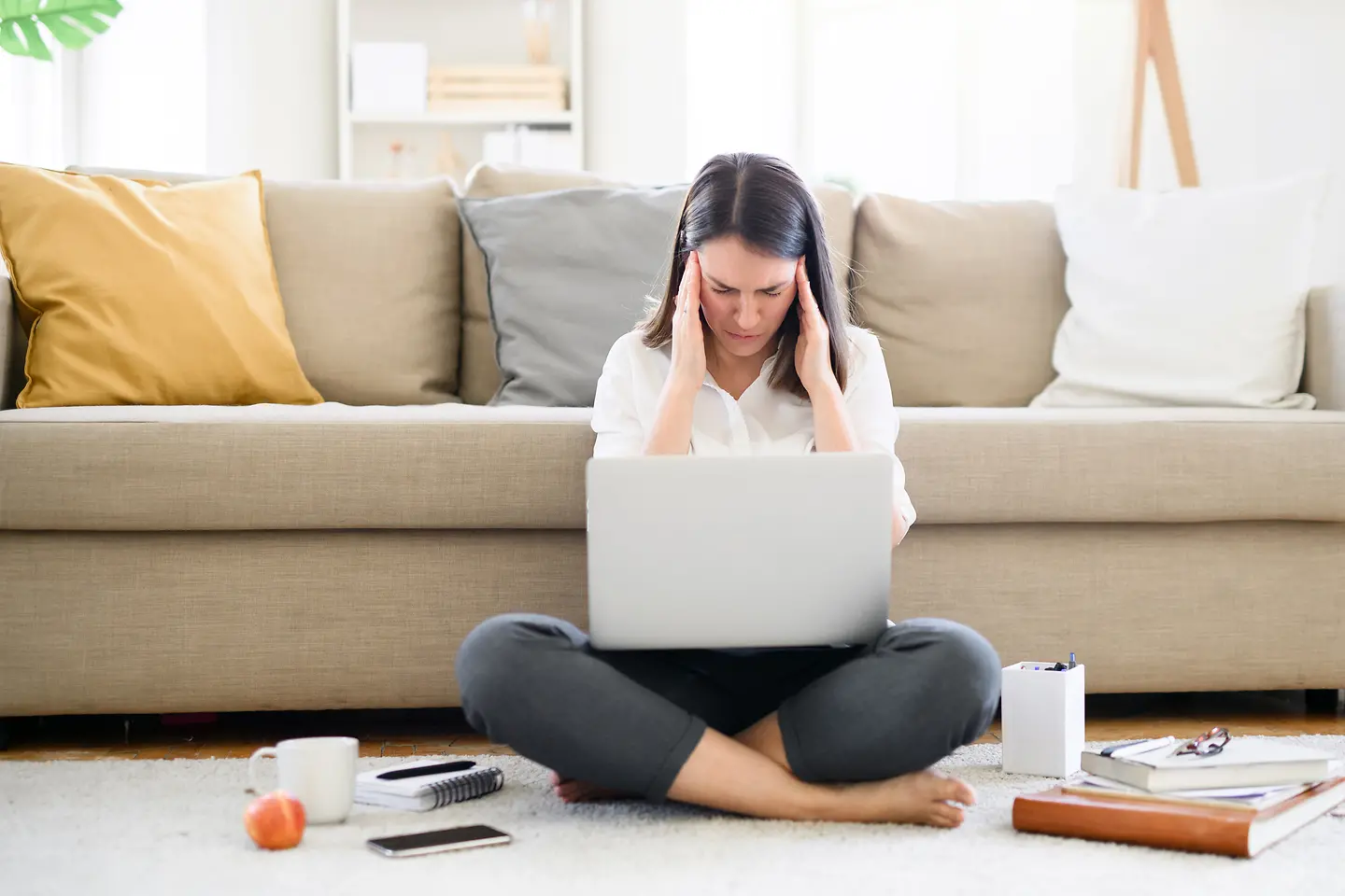Frau sitzt mit Arbeitsutensilien und Kopfschmerzen auf dem Wohnzimmerfussboden