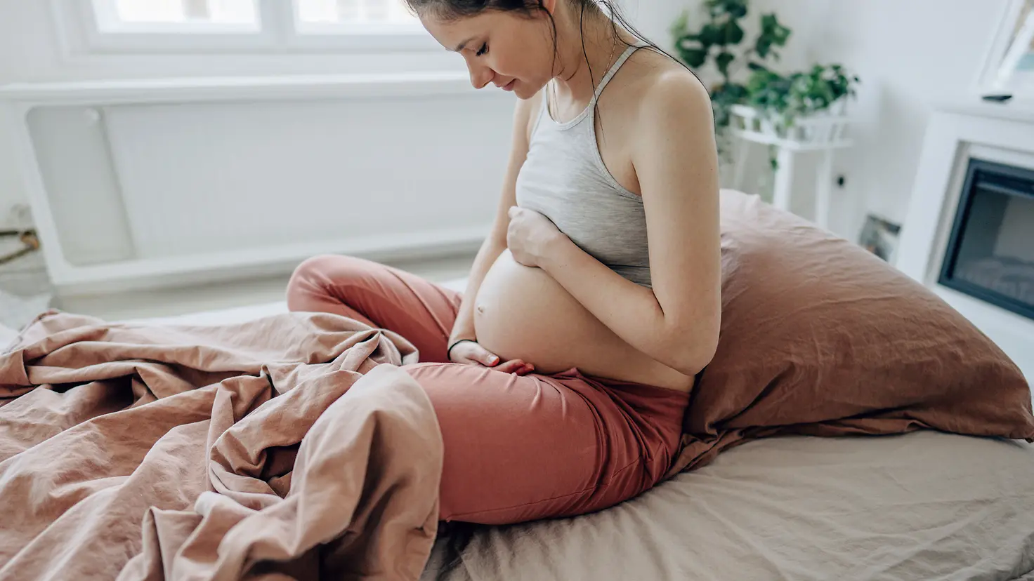 Eine junge schwangere Frau um die 31. Schwangerschaftswoche sitzt im Bett und streichelt ihren Babybauch