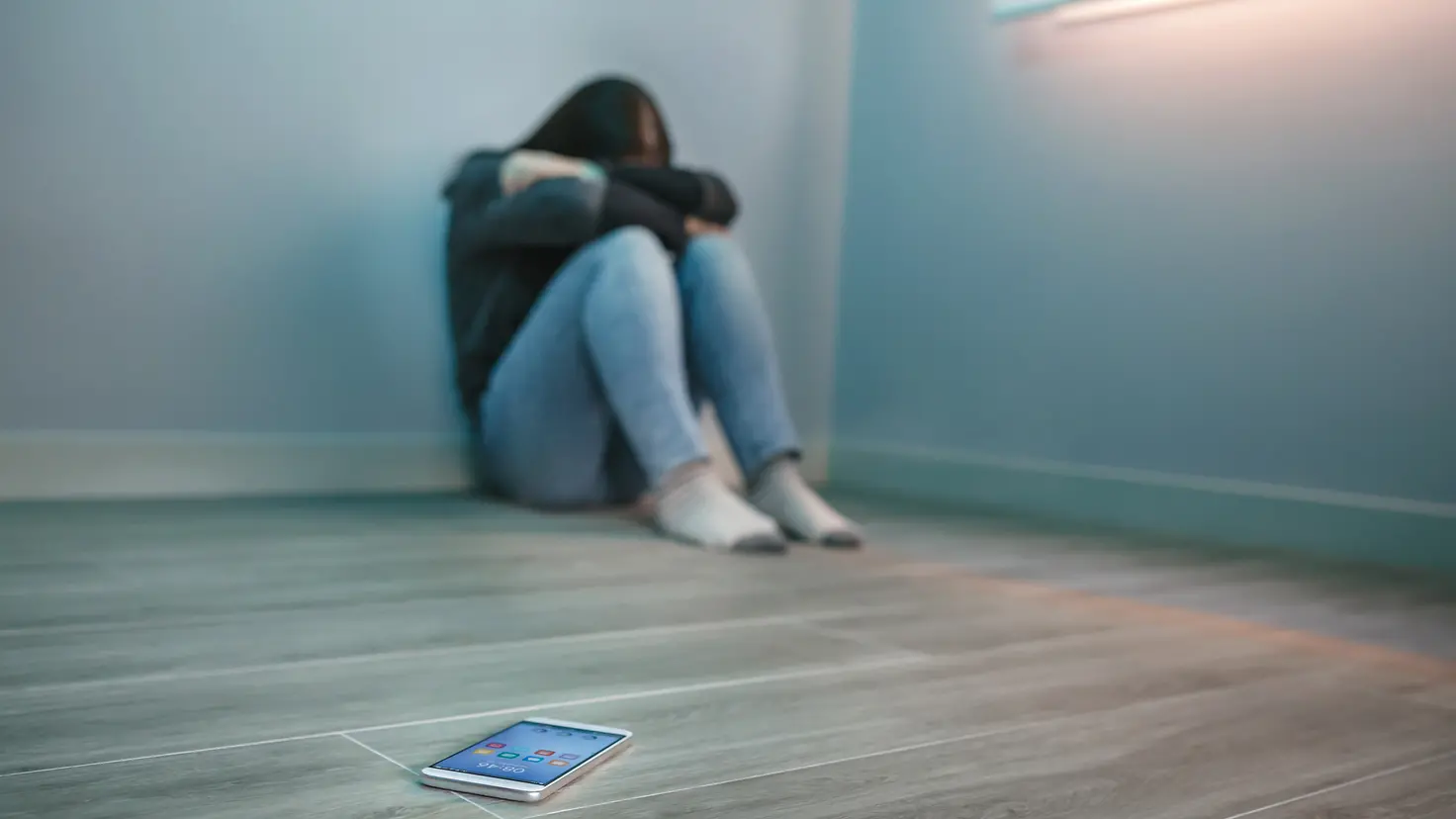 Zu sehen ist eine auf dem Boden sitzende junge Frau mit verschränkten Armen vor dem Gesicht und ein Smartphone