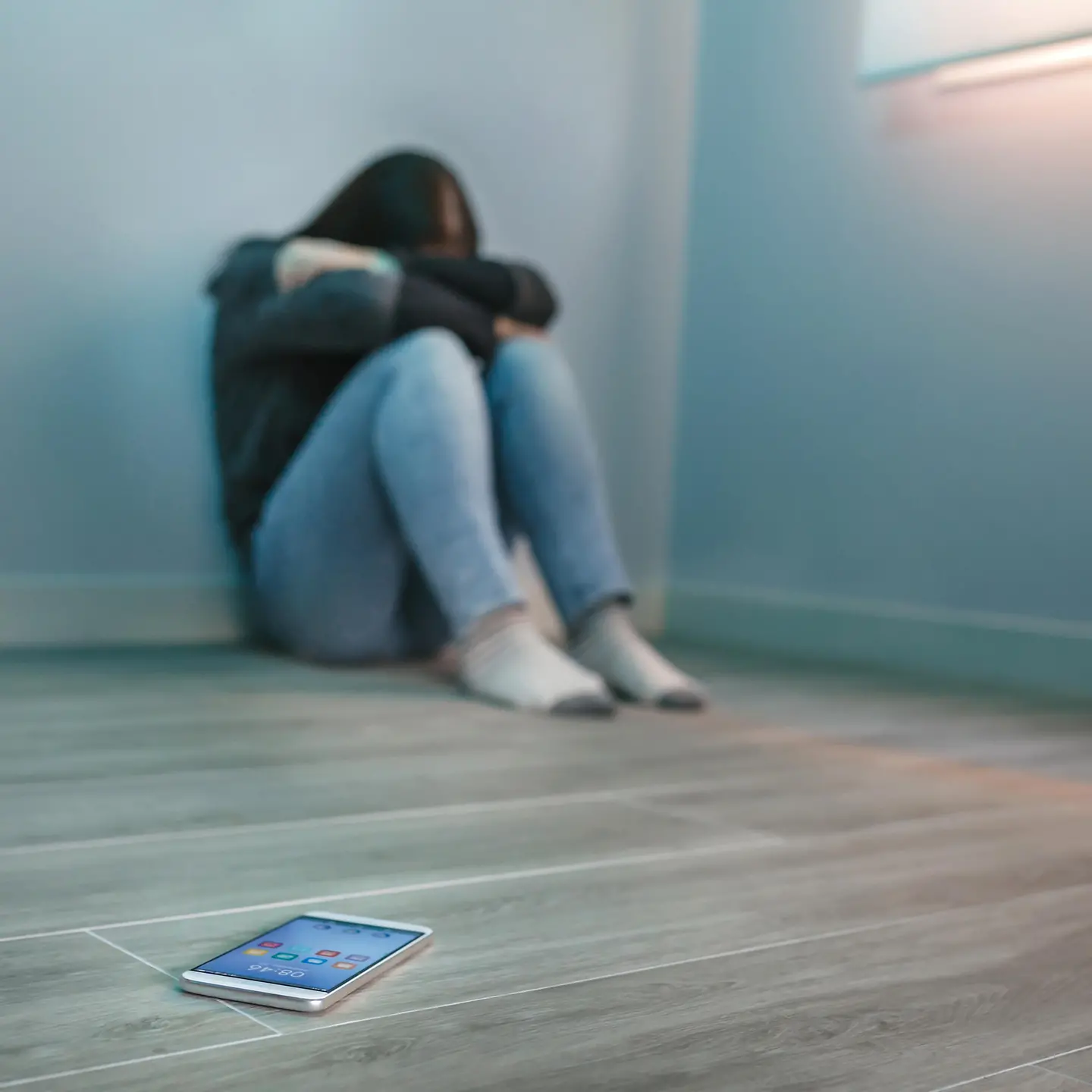 Zu sehen ist eine auf dem Boden sitzende junge Frau mit verschränkten Armen vor dem Gesicht und ein Smartphone