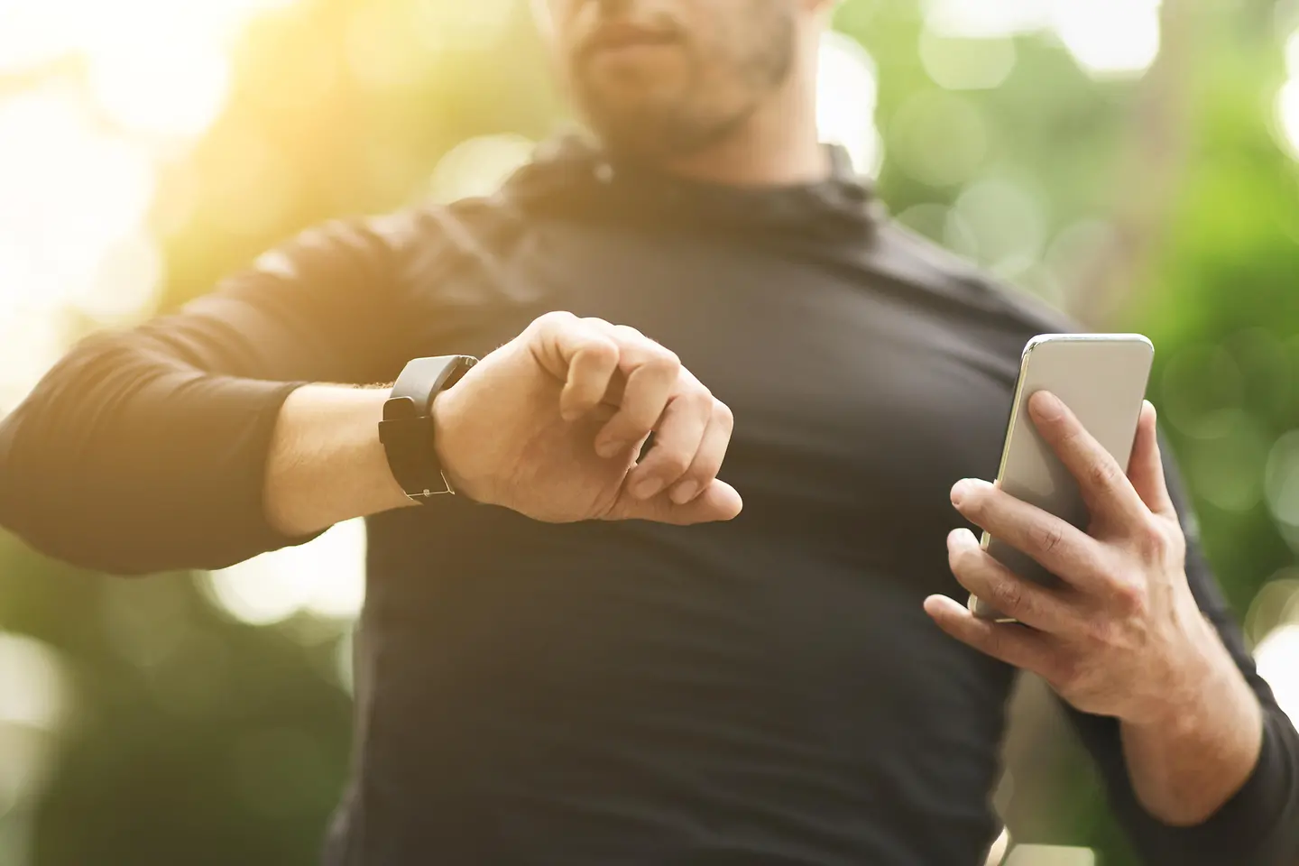 Zu sehen ist ein Mann, der auf seine Fitnessuhr sieht und in der anderen Hand ein Smartphone hält