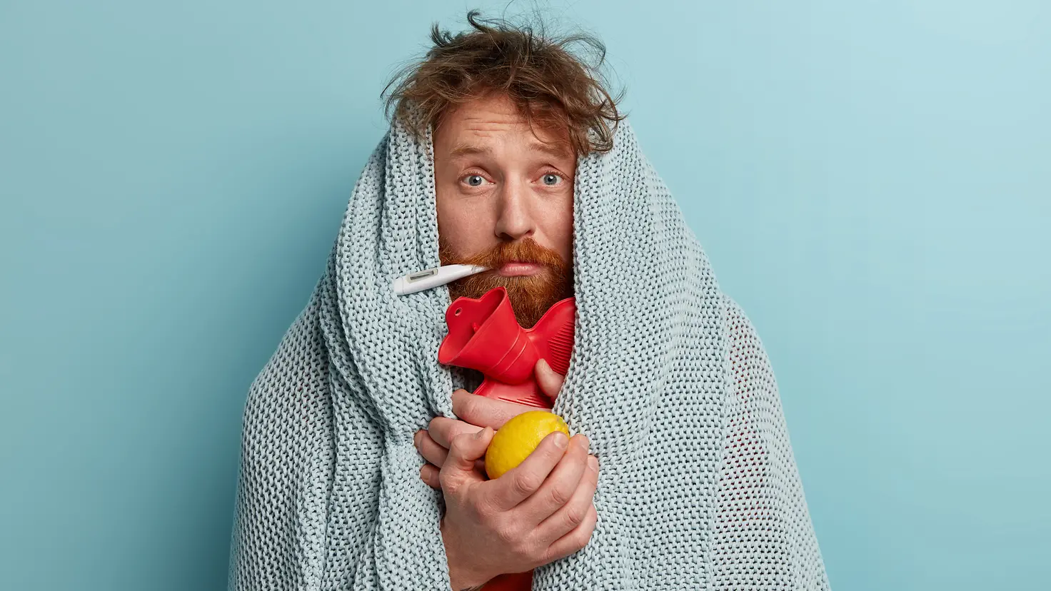 Zu sehen ist ein Mann, eingewickelt in eine Decke und mit Fieberthermometer, Wärmflasche und einer Zitrone