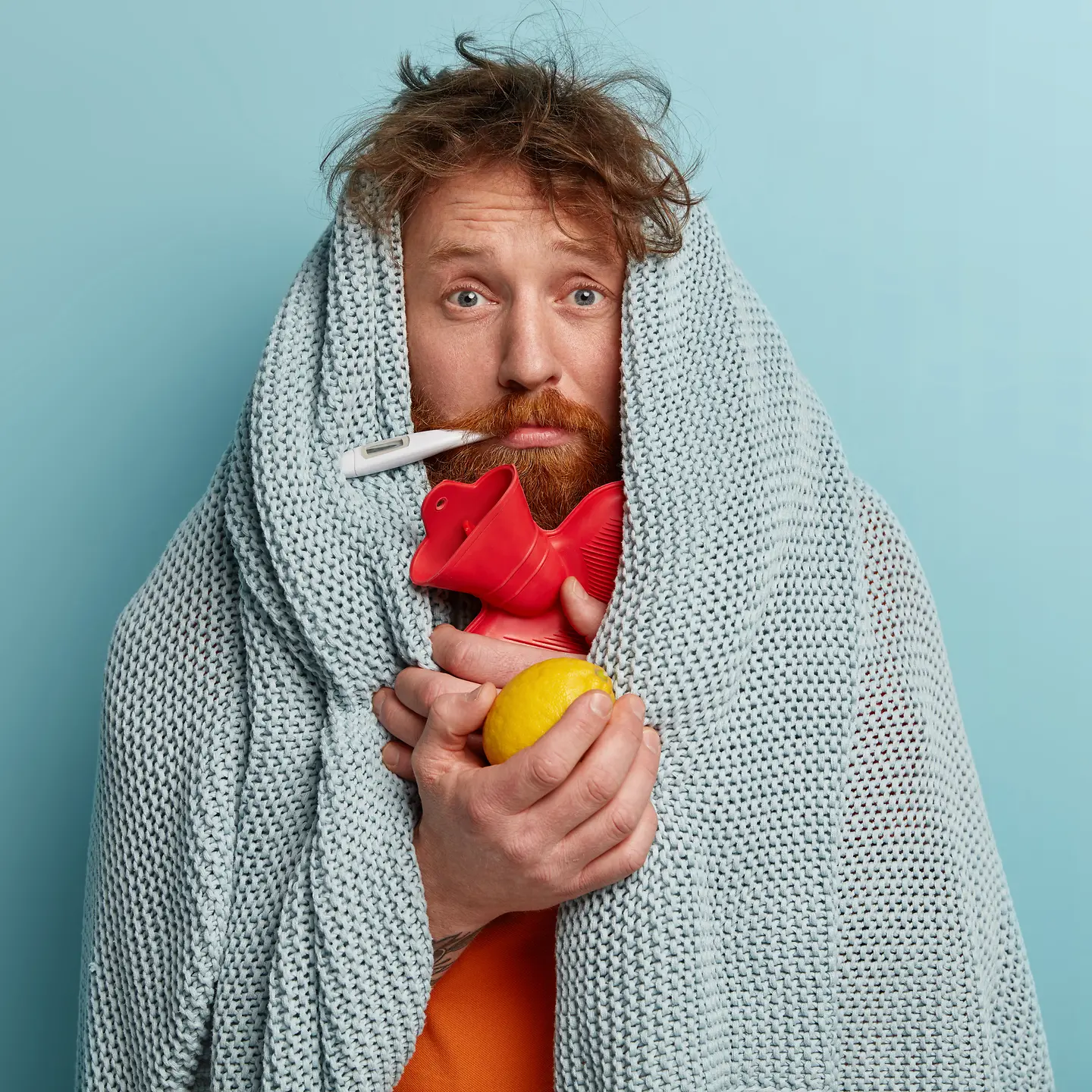Zu sehen ist ein Mann, eingewickelt in eine Decke und mit Fieberthermometer, Wärmflasche und einer Zitrone