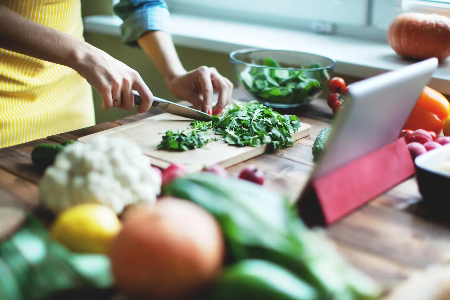 Eine Person (einen Kopf sieht man nicht) schneidet frisches Gemüse und Kräuter auf einem Holzbrett auf einem Tisch.