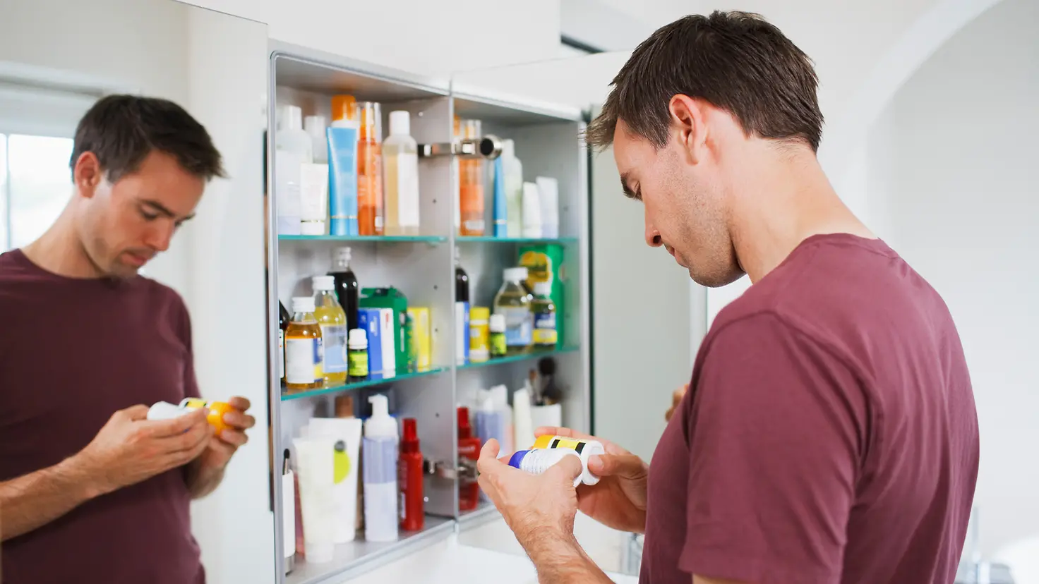 Ein Mann guckt skeptisch auf zwei Medikamentendosen im Badezimmer