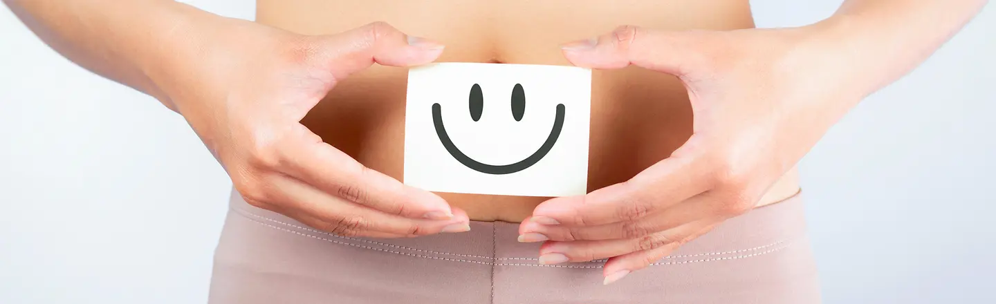 Eine Frau hält einen Zettel vor ihren Bauch. Auf dem Zettel ist ein positiver Smiley abgebildet.