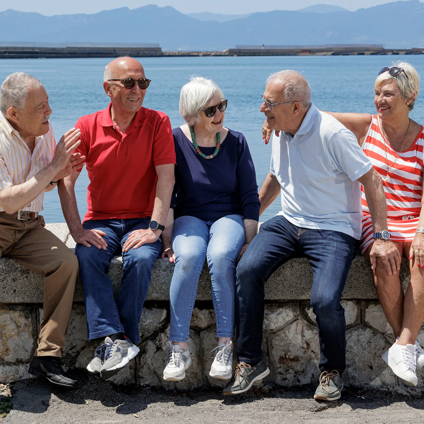 Zu sehen sind fünf ältere Personen, die auf einer Mauer am Meer sitzen