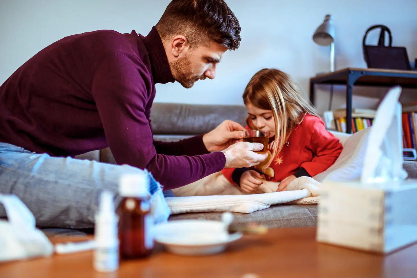 Zu sehen ist ein Vater, der seiner kleinen kranken Tochter Tee zum trinken gibt