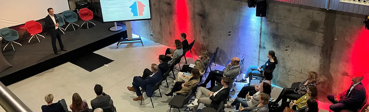 Blick von oben in eine Halle, dort steht ein Mann auf einer Bühne und hält einen Vortrag.