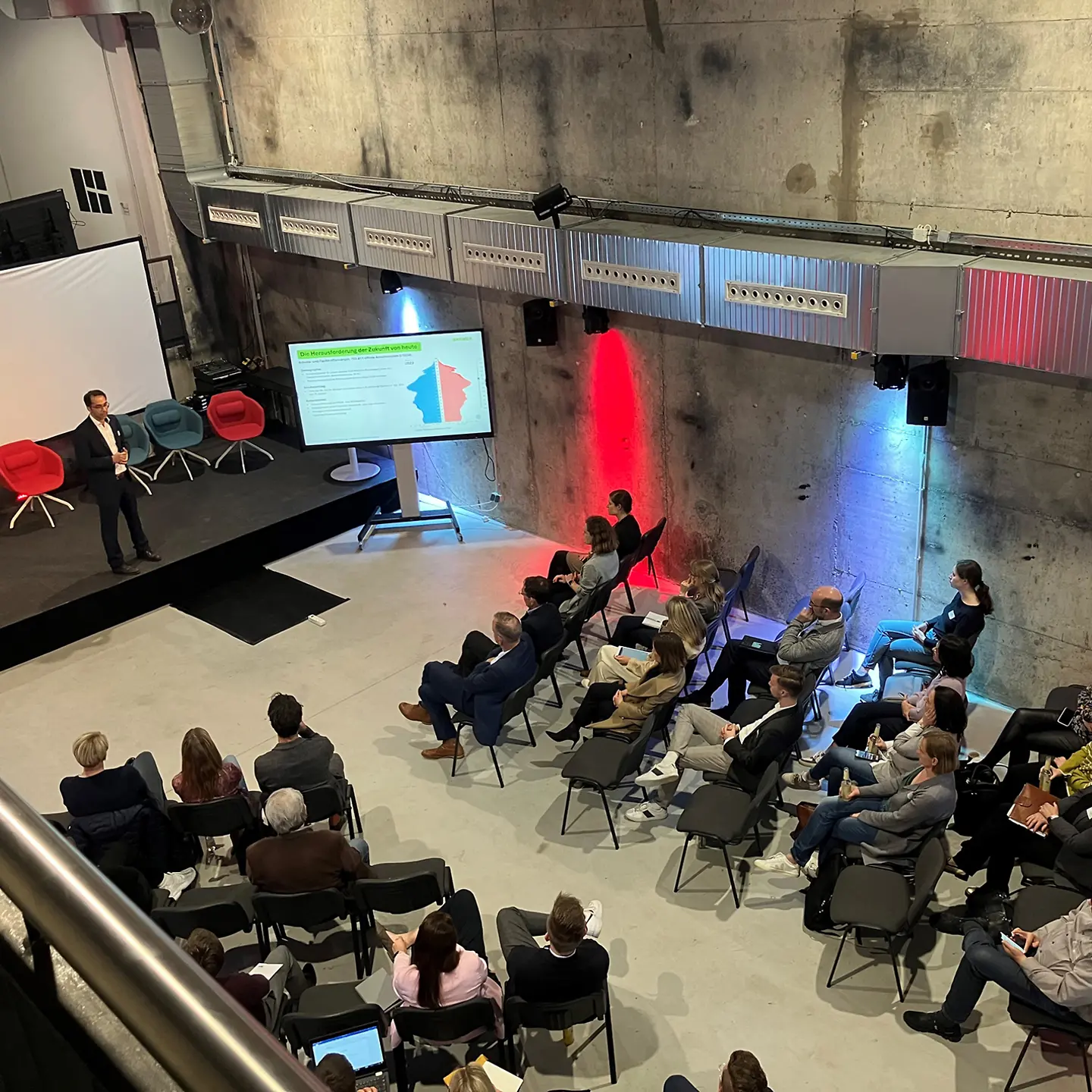 Blick von oben in eine Halle, dort steht ein Mann auf einer Bühne und hält einen Vortrag.