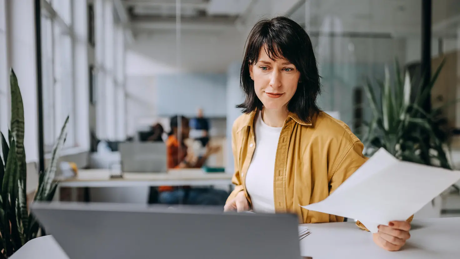 Erwachsene Frau in modernem Büro schaut auf den Laptop und sortiert Dokumente