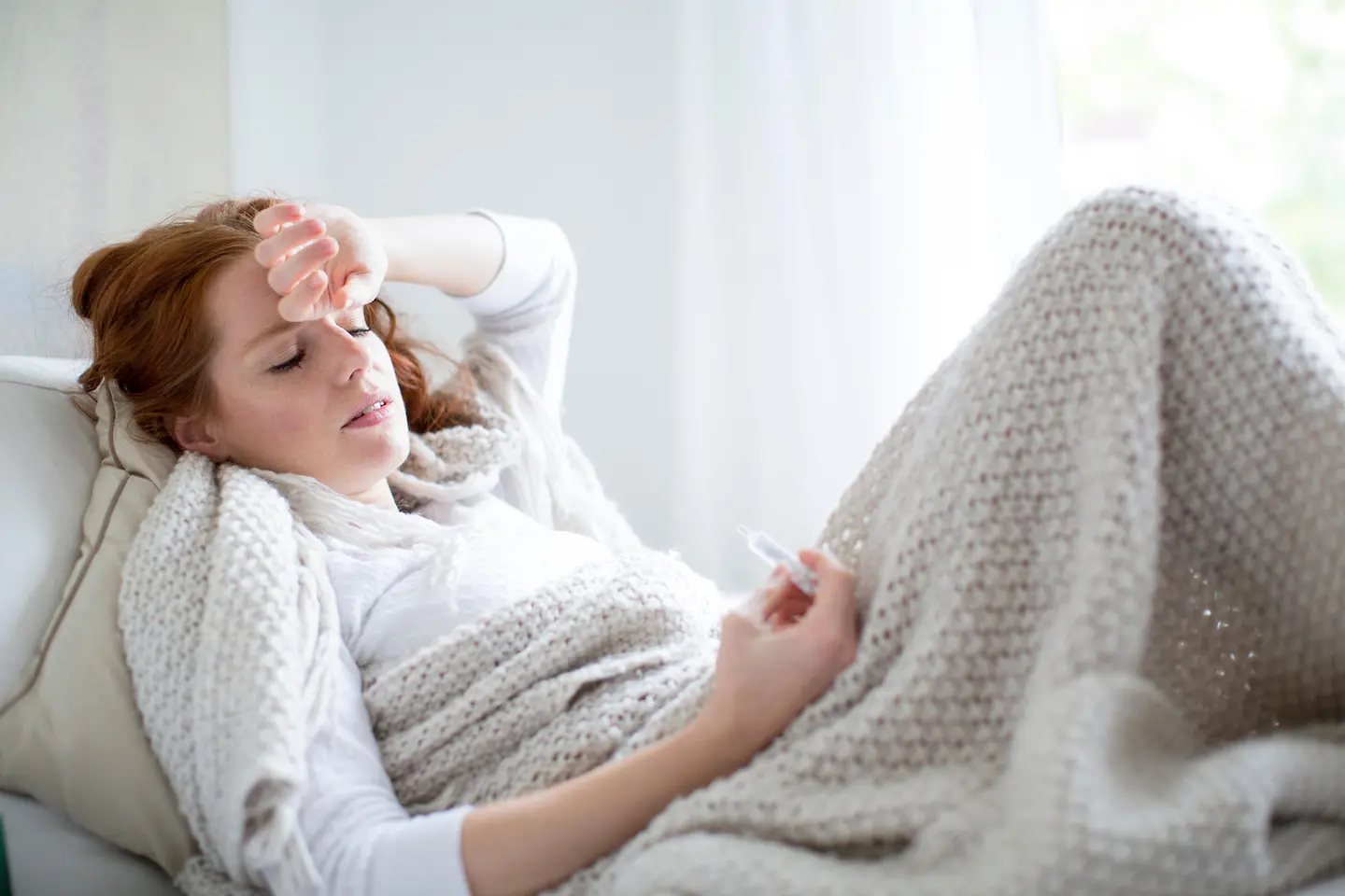 Zu sehen ist eine Frau unter einer wärmenden Decke mit Fieberthermometer in der Hand