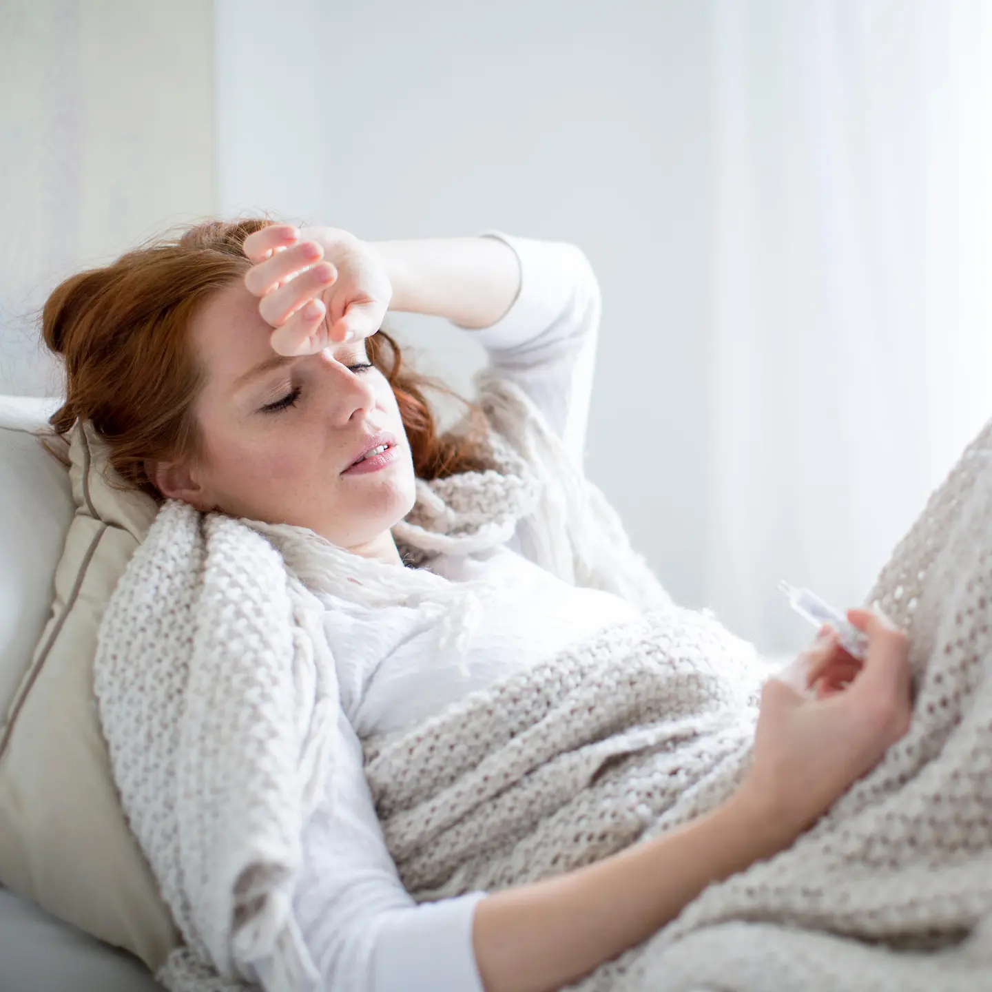 Zu sehen ist eine Frau unter einer wärmenden Decke mit Fieberthermometer in der Hand
