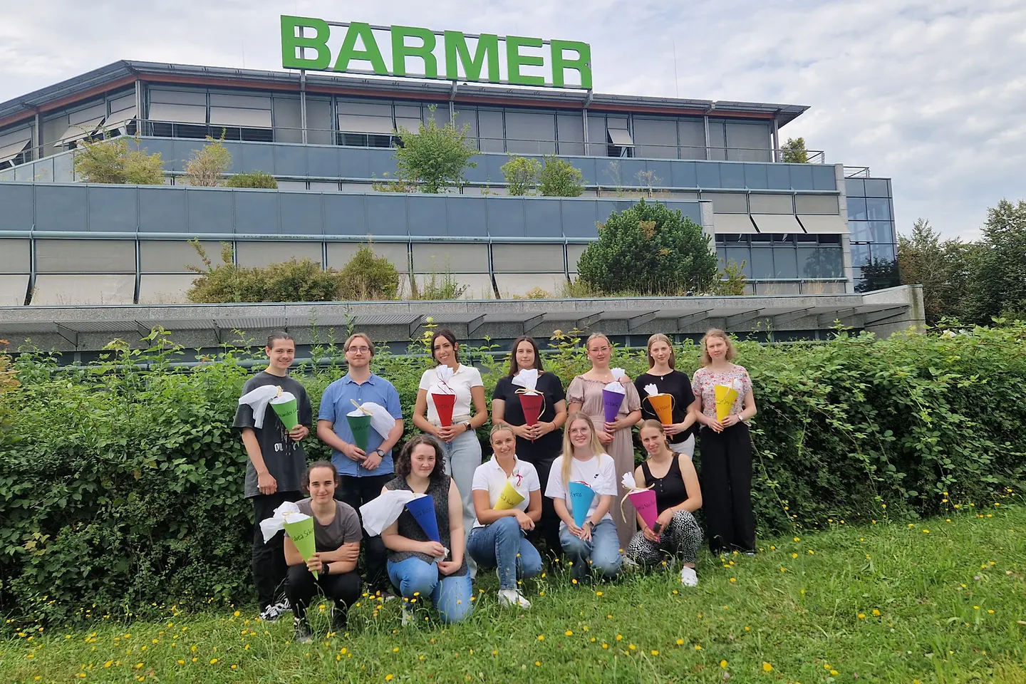 Zwölf junge Frauen und Männer mit Schultüten in der Hand stehen und hocken vor einem Bürogebäude mit der Aufrschrift 'BARMER'