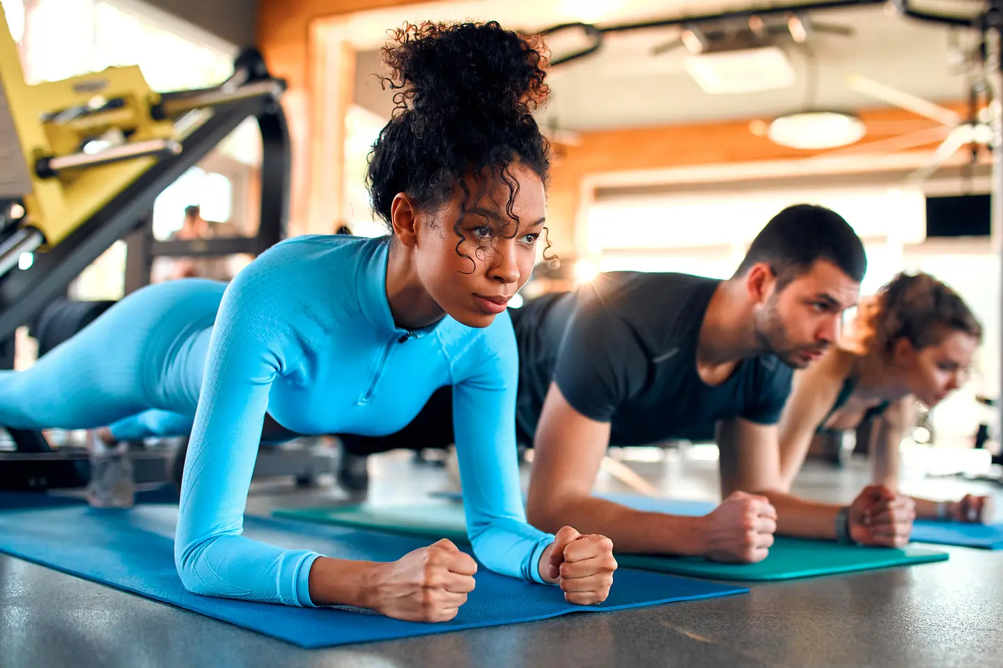 Zu sehen auf diesem Bild sind zwei Frauen und ein Mann. Sie befinden sich alle drei nebeneinander auf Turnmatten in der Eigengewichtsübung des Unterarmstützes, auch bekannt als Plank. 