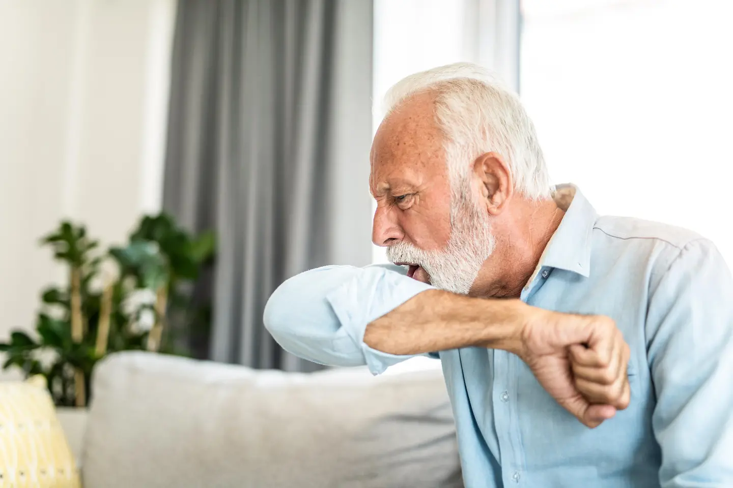 Ein Mann im höheren Alter sitzt auf einem Sofa und hustet in seine Armbeuge