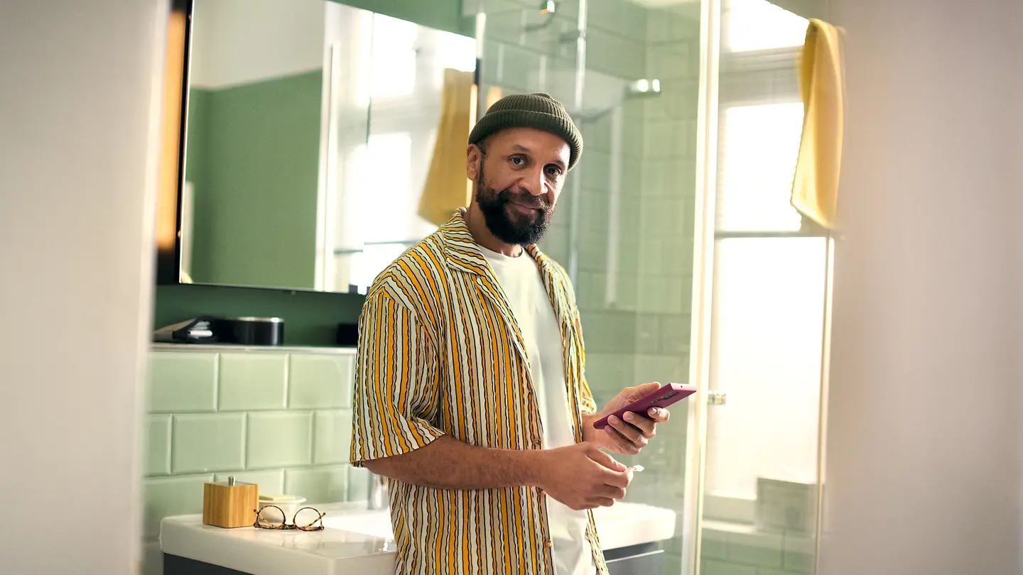 Ein Mann steht im Badezimmer und hält sein Smartphone und ein Medikament in der Hand