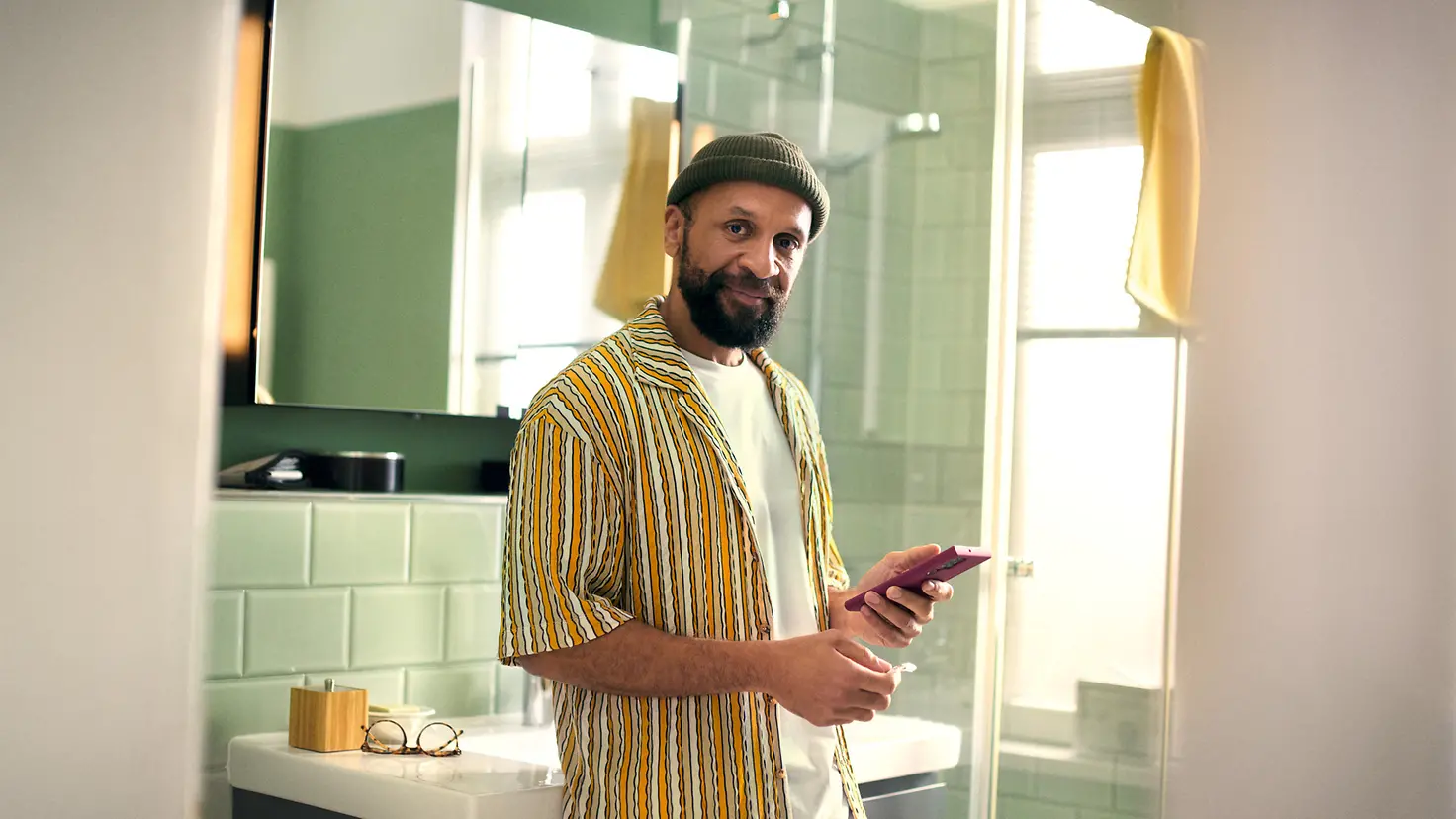 Ein Mann steht im Badezimmer und hält sein Smartphone und ein Medikament in der Hand
