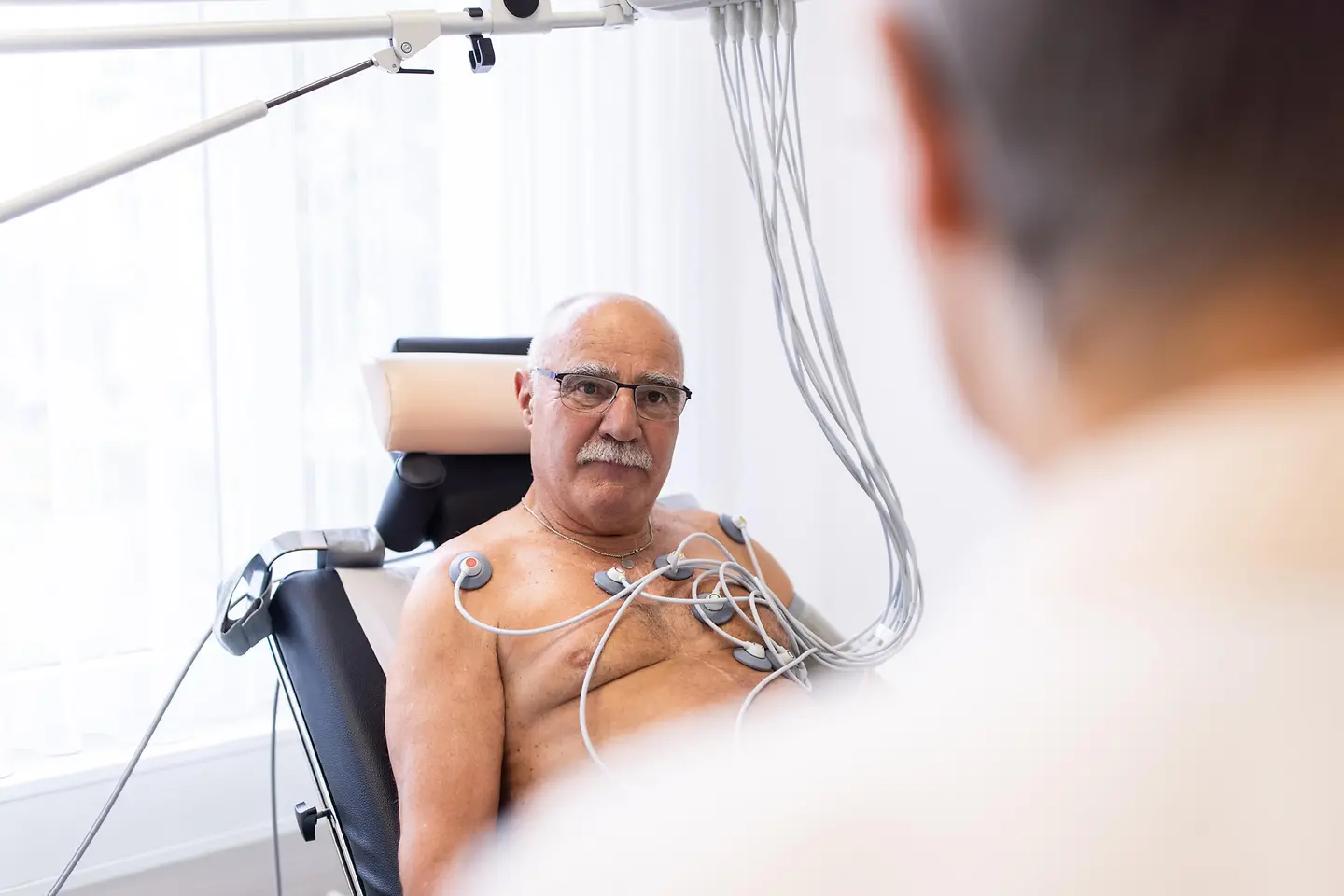Älterer männlicher Patient mit EKG-Sensor auf der Brust schaut einen Arzt in der Klinik an. Der Senior führt einen Herzbelastungstest in der kardiologischen Klinik durch.