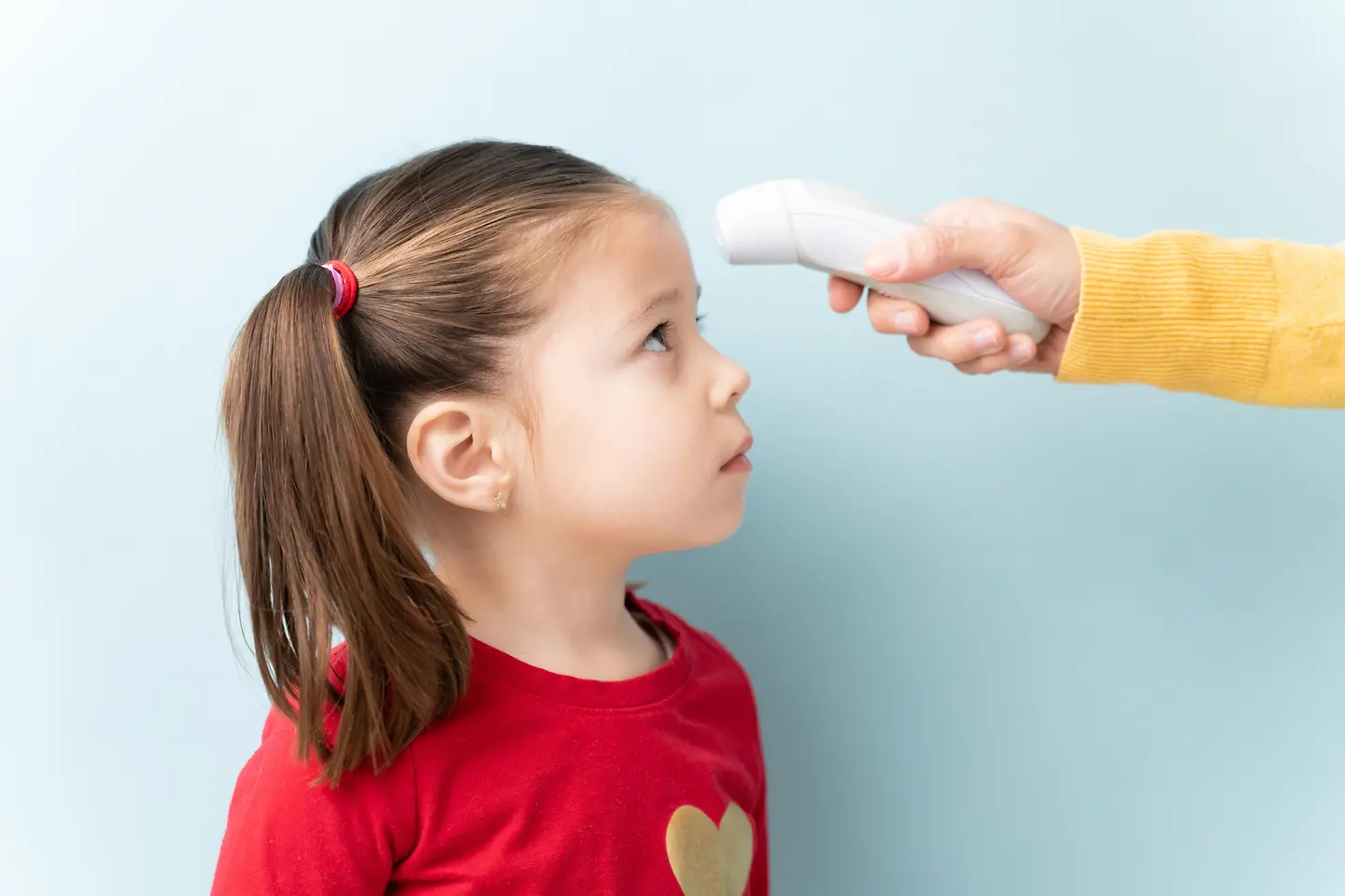 Ein kleines Mädchen mit braunen Haaren, die zu einem seitlichen Pferdeschwanz gebunden sind, trägt ein rotes Shirt mit einem goldenen Herz darauf. Eine Person misst ihre Temperatur mit einem Stirnthermometer. 