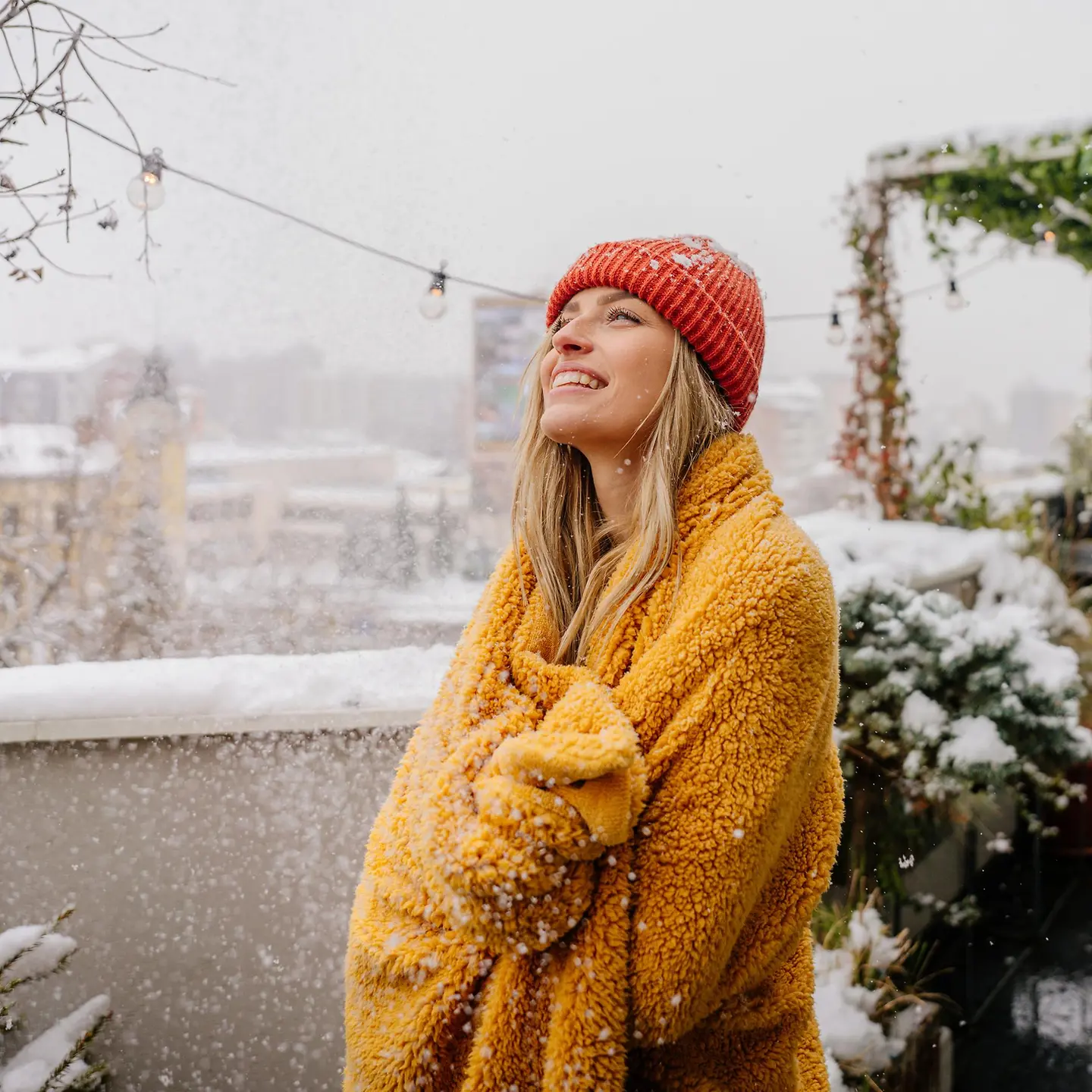 Eine junge Frau steht im Schnee auf einem Balkon in eine Decke eingewickelt