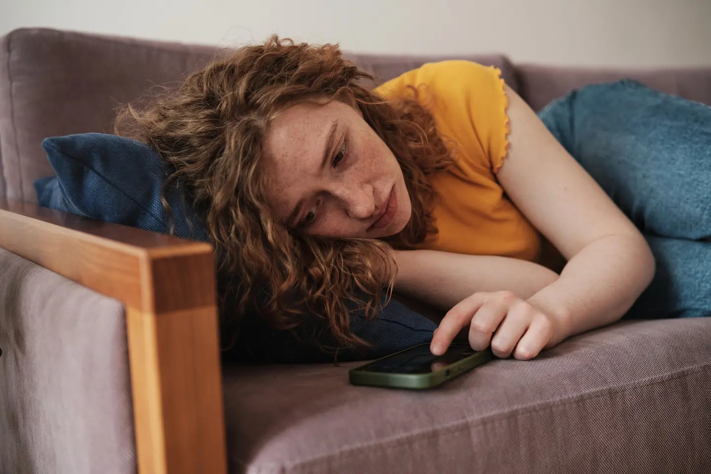 Junge Frau mit roten Haaren liegt auf einer Couch und schaut auf ihr Handy