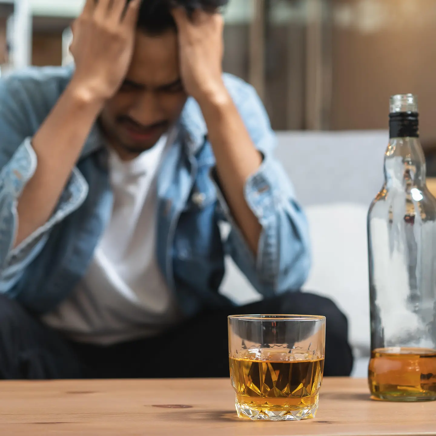 Zu sehen ist ein Mann mit seinen Händen am Kopf vor einer Flasche und einem Glas Alkohol