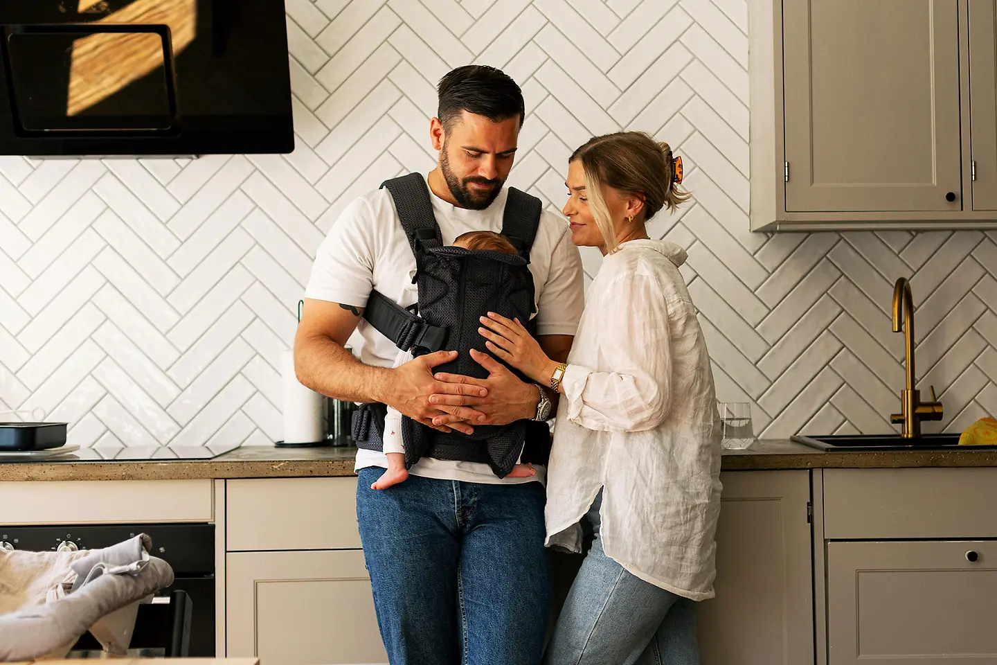 Ein junges Eltern-Paar, mit Baby in einer Babytrage am Bauch, steht in einer Küche.