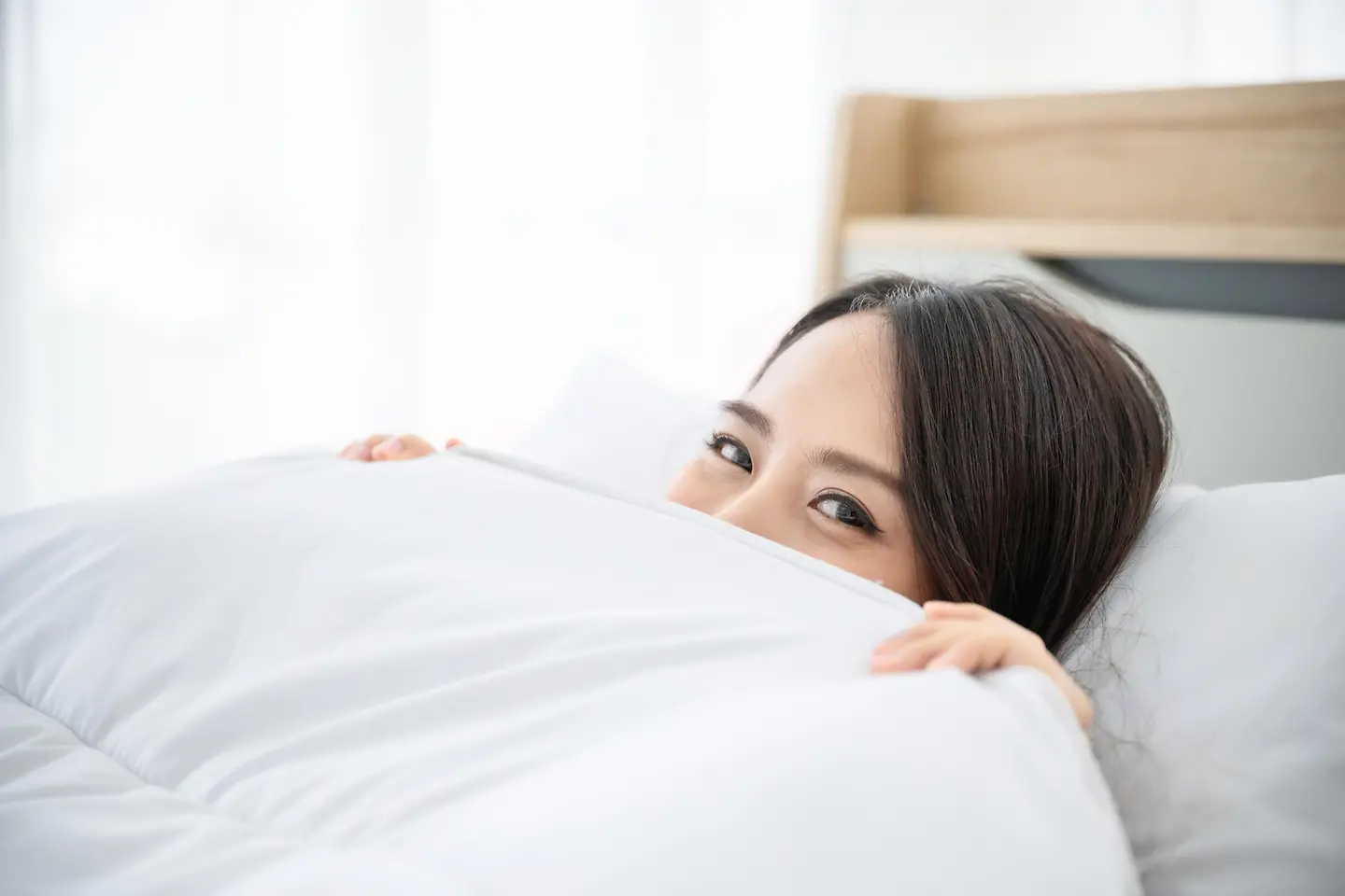 Zu sehen auf diesem Bild ist eine Frau, die unter einer Bettdecke im Bett liegt. Von ihr zu sehen sind die beiden Hände, die die Bettdecke festhalten, sowie die obere Hälfte ihres Gesichts ab Nase.