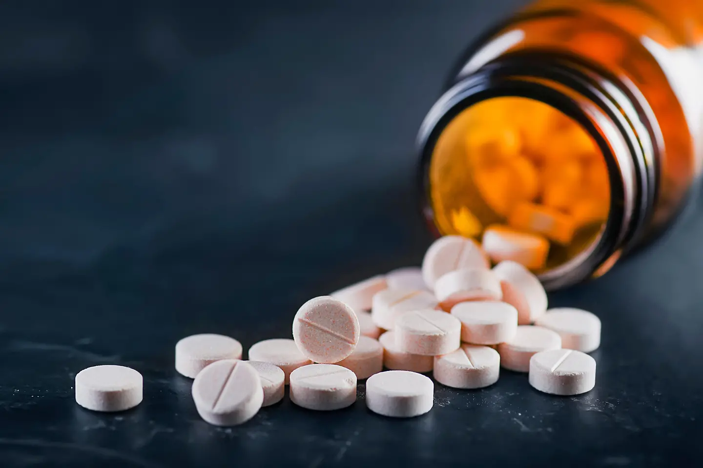 Zu sehen auf diesem Bild ist ein auf der Seite liegendes offenes Fläschchen zu sehen. Die in dem Fläschchen enthaltenden Pillen sind herausgefallen und liegen vor dem Fläschchen.