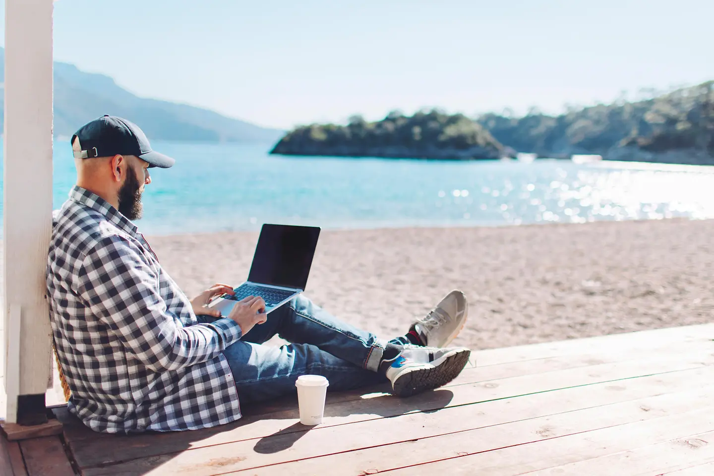 Junger Mann mit Basecap arbeitet am Strand mit dem Laptop