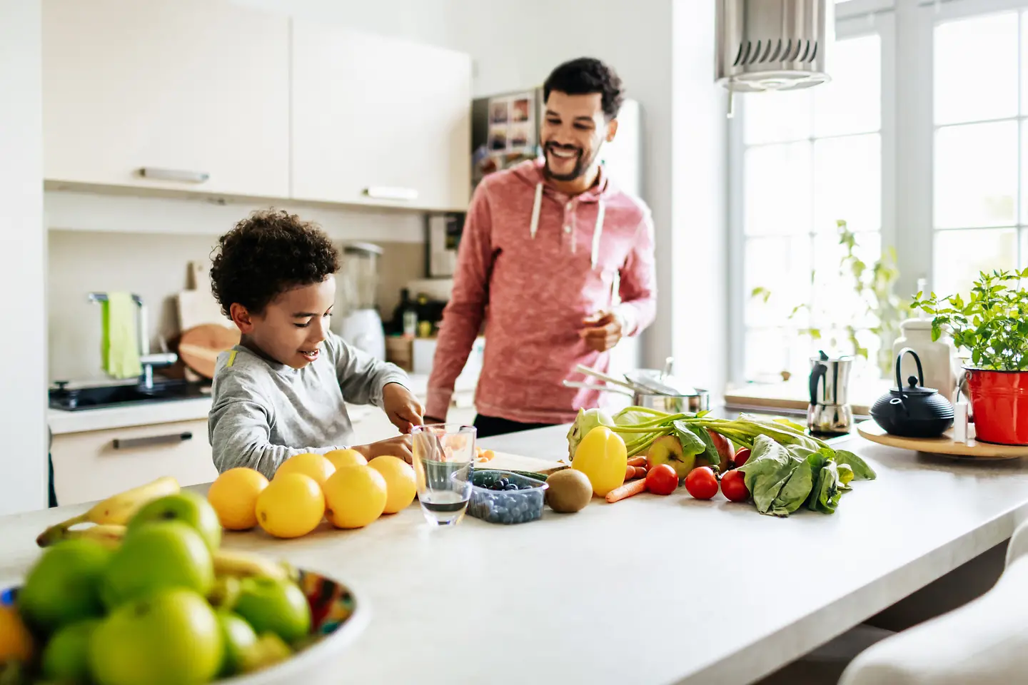 Ein Junge schneidet Obst und Gemüse auf einer Arbeitsplatte in einer Küche. Sein Dad steht daneben und lächelt.