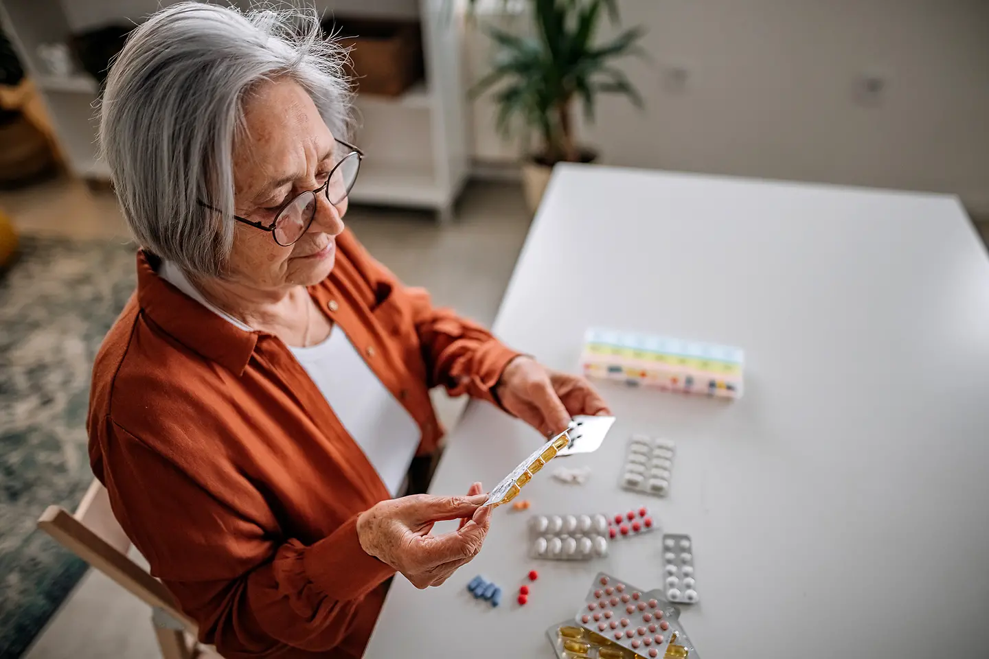 Eine Seniorin sitzt an einem Tisch mit vielen Medikament-Packungen. Sie hält und betrachtet verschiedene Medikamente.