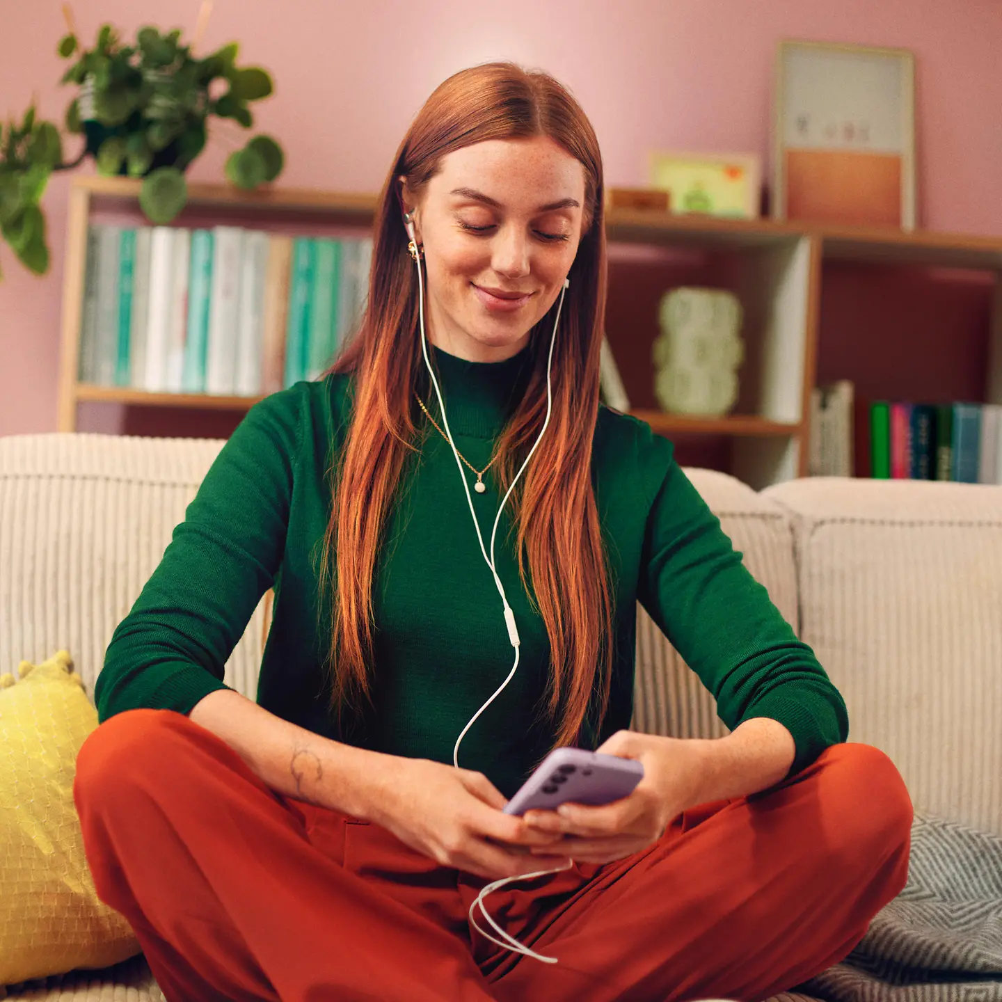 A woman sits on her sofa and listens to music on her cell phone through headphones.