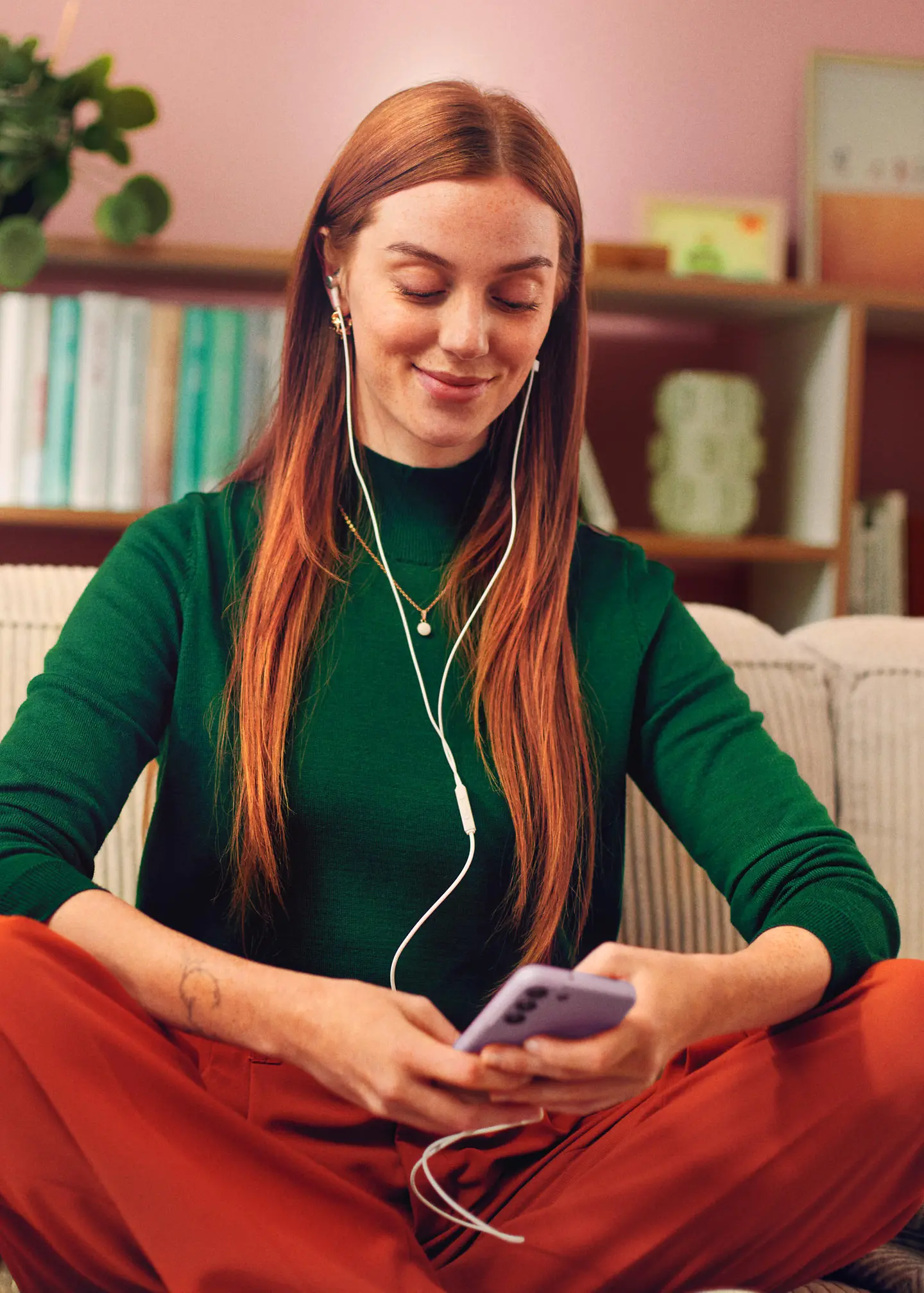 A woman sits on her sofa and listens to music on her cell phone through headphones.