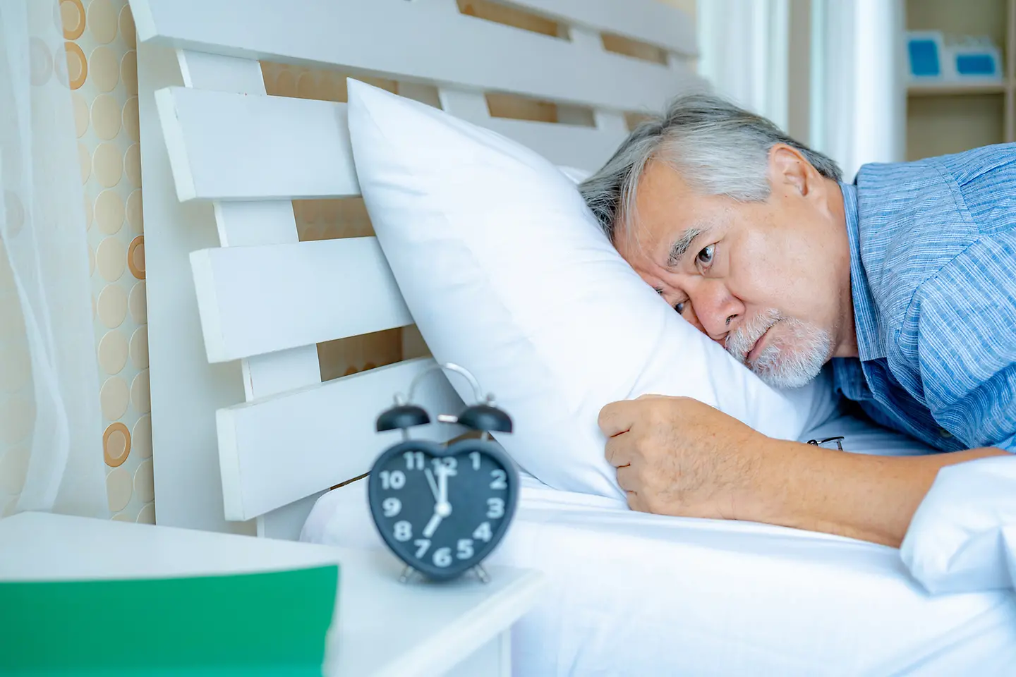 Zu sehen auf dem Bild ist ein älterer Mann, der im mit offenen Augen im Bett liegt und in Richtung des Weckers auf dem Nachtschrank blickt. 