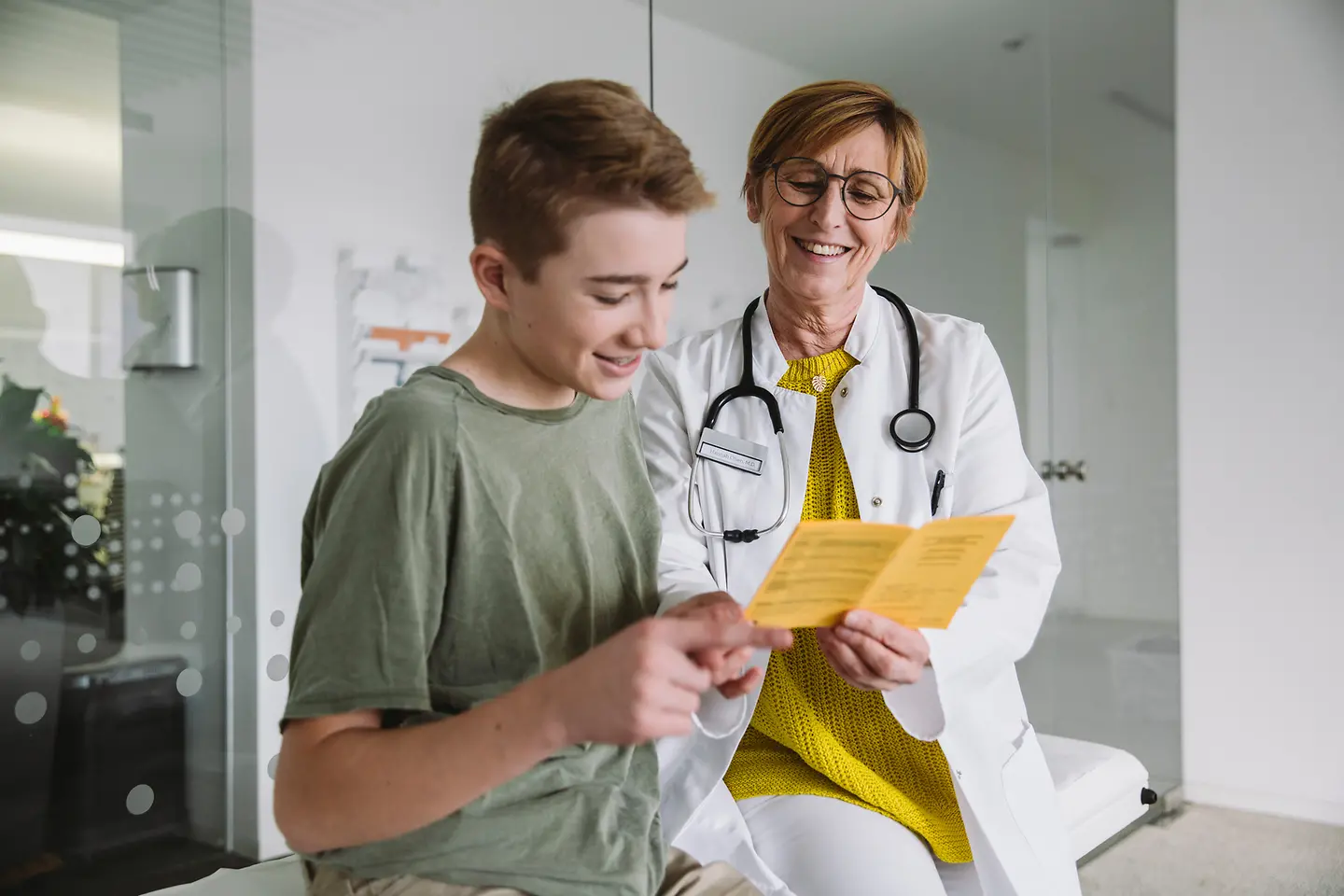 Ein junger Mann sieht sich gemeinsam mit einer Ärztin einen Impfausweis an. Beide lächeln. 