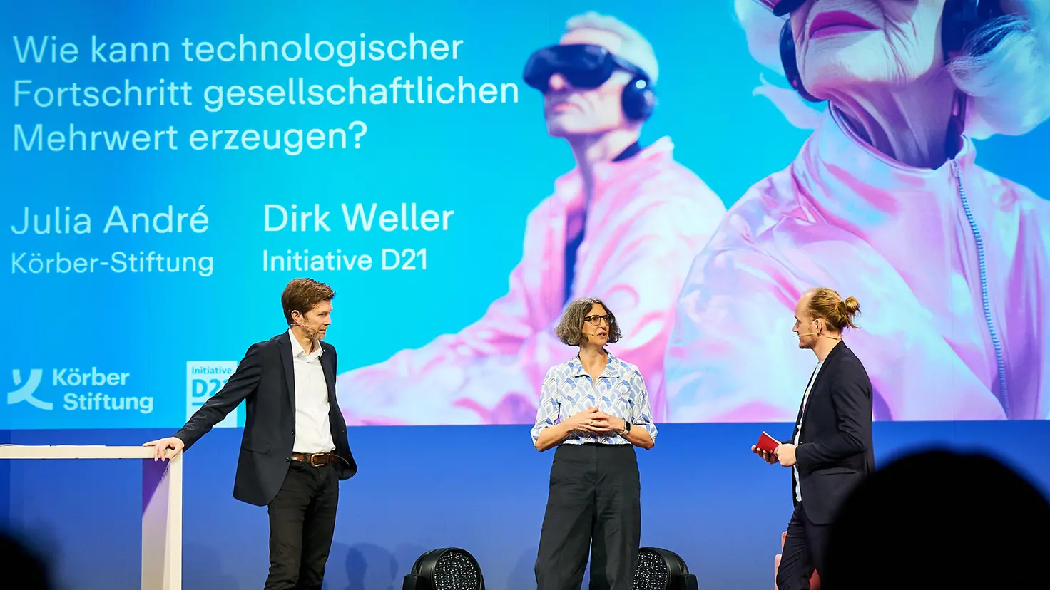 Drei Moderatoren stehen auf einer Bühne und diskutieren. Dahinter eine Leinwand mit dem Slogan "Technologie im Alter: Mehr als Sturzmatte & Seniorenhandy"