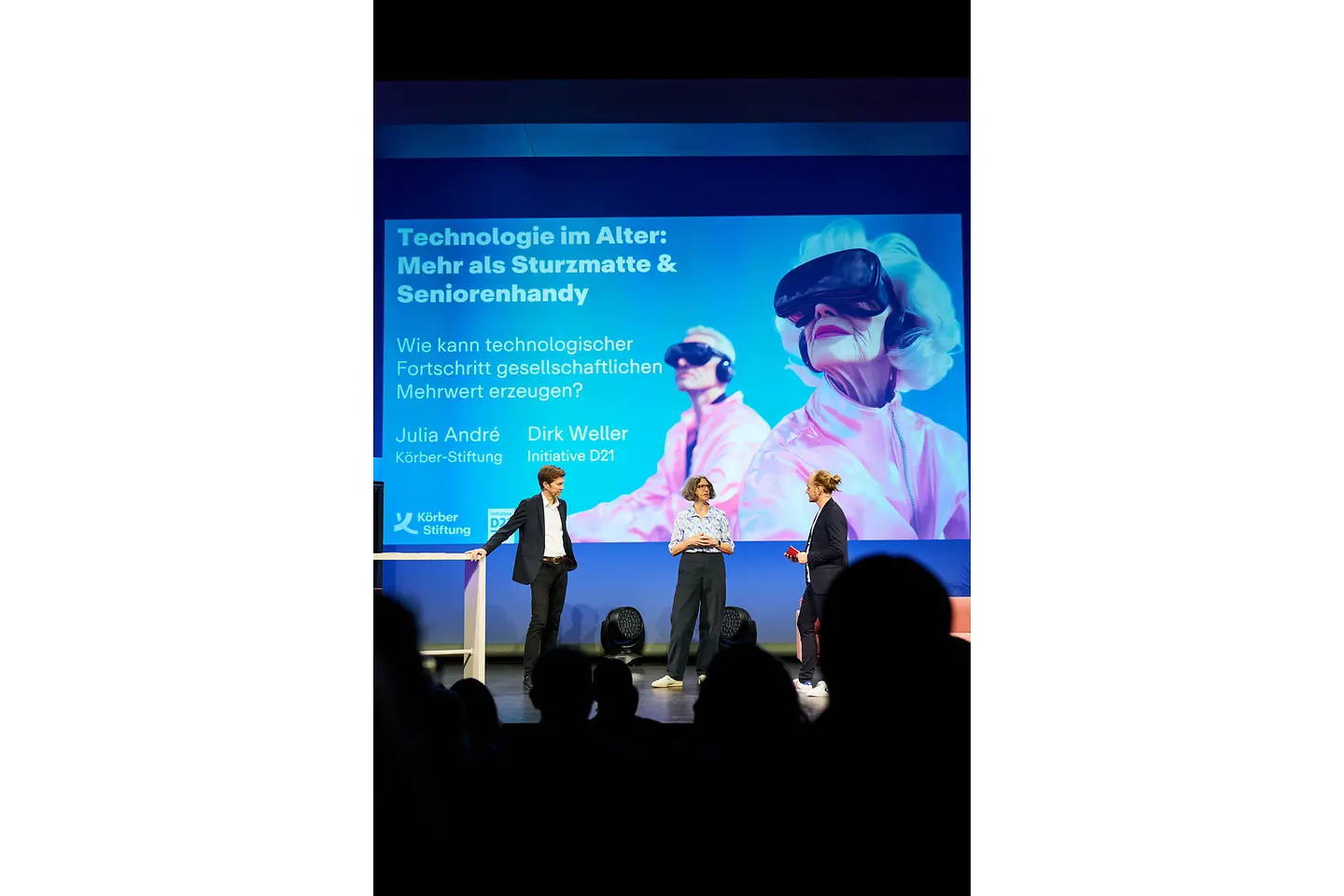 Drei Moderatoren stehen auf einer Bühne und diskutieren. Dahinter eine Leinwand mit dem Slogan "Technologie im Alter: Mehr als Sturzmatte & Seniorenhandy"
