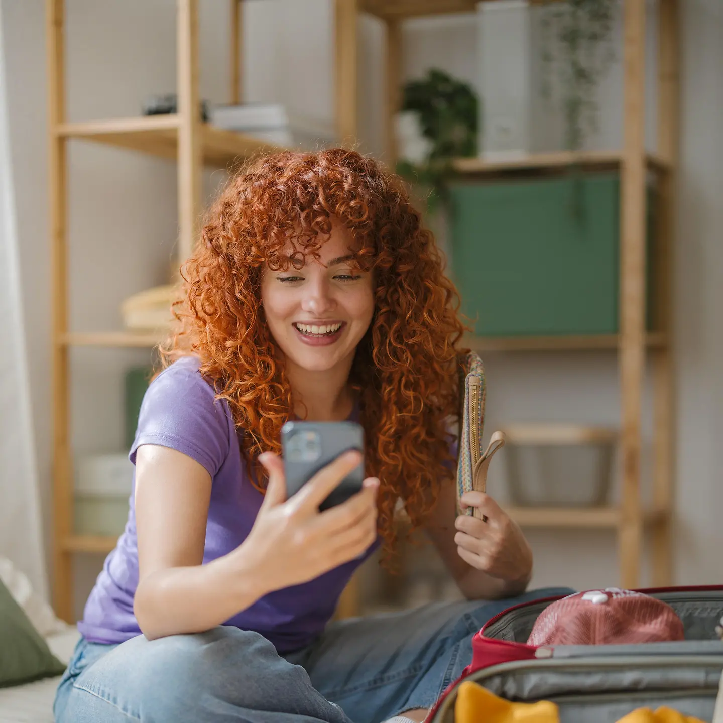 Frau, die auf dem Bett sitzt, ihren Koffer packt und dabei mit dem Smartphone über Videotelefonie telefoniert.