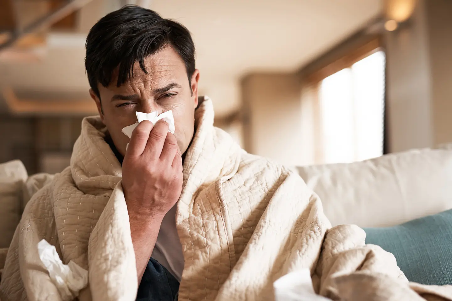 Ein erkälteter Mann sitzt mit umgelegter Decke auf dem Sofa und schnäuzt sich die Nase.
