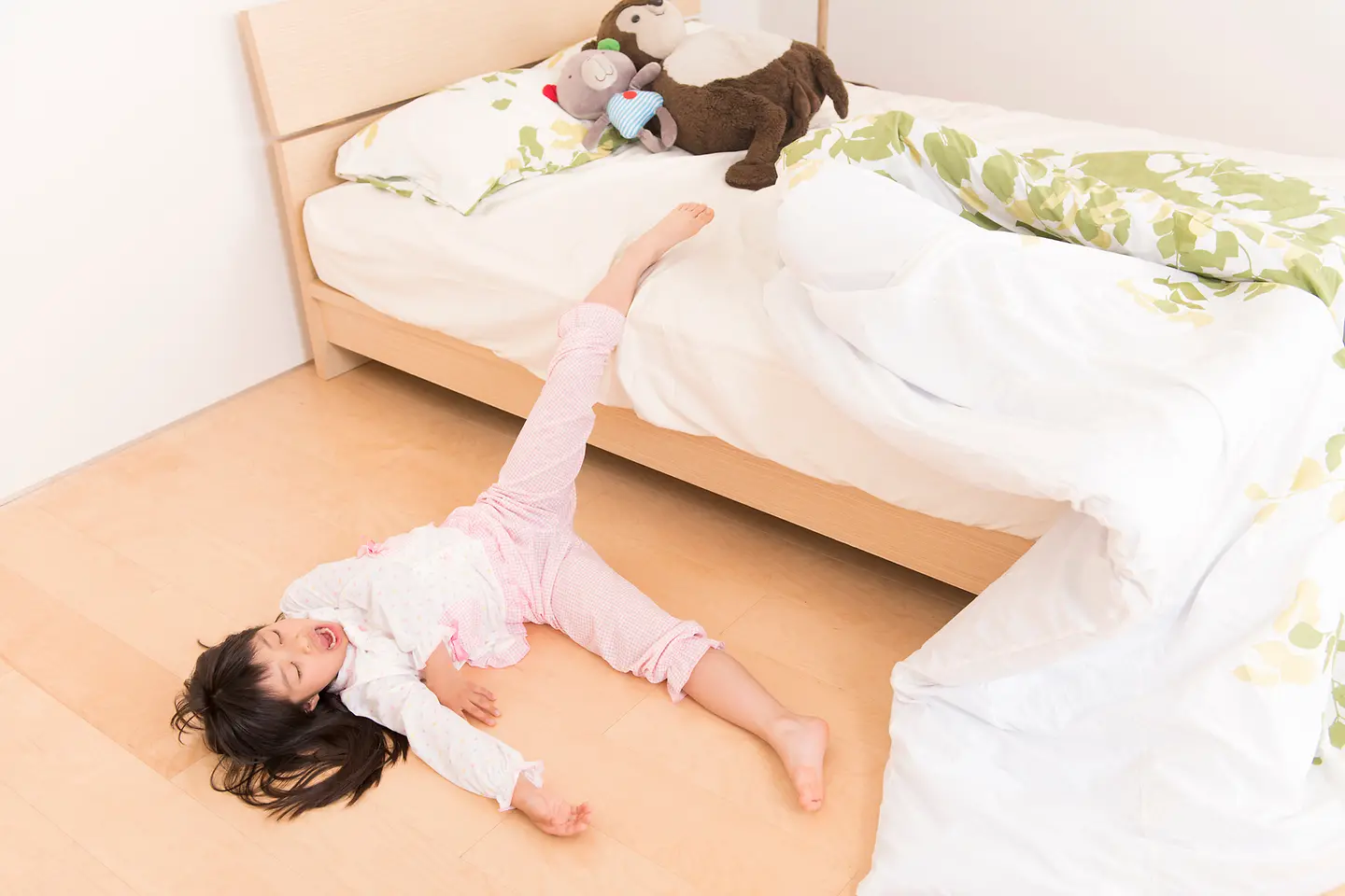 Auf diesem Bild ist ein Kind zu sehen, dass scheinbar aus dem Bett gefallen ist. Zumindest liegt es schlafend auf dem Fußboden, während das linke Bein nach oben ausgestreckt ist und noch Kontakt zum Kinderbett hat.