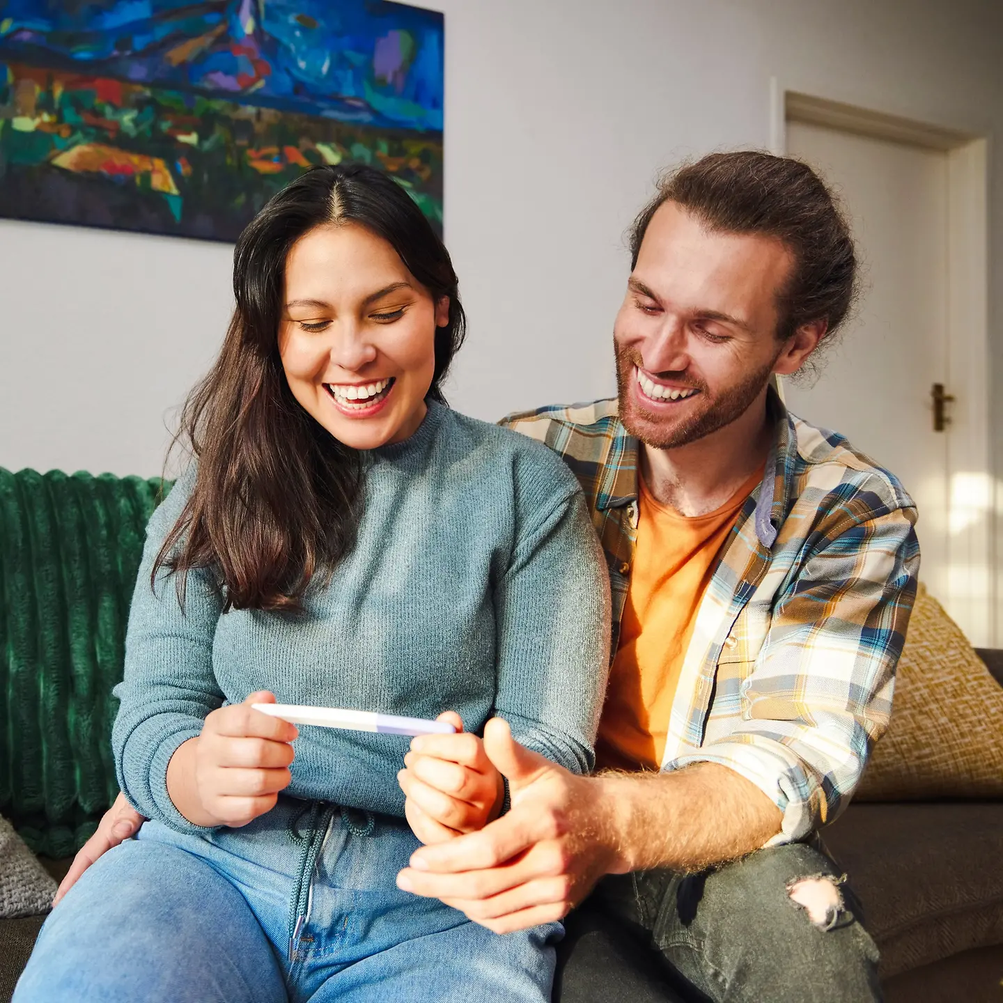 Ein junges Paar sitzt zusammen auf einem Sofa und schaut glücklich lachend auf einen Schwangerschaftstest.